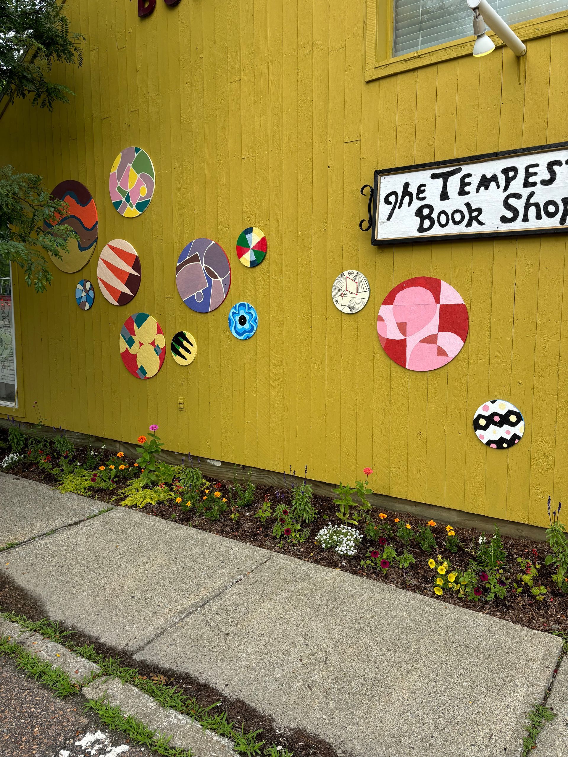 Colorful paper circles on a yellow wall beside a sidewalk and a handmade sign reading “St. Tempes Book Shop”