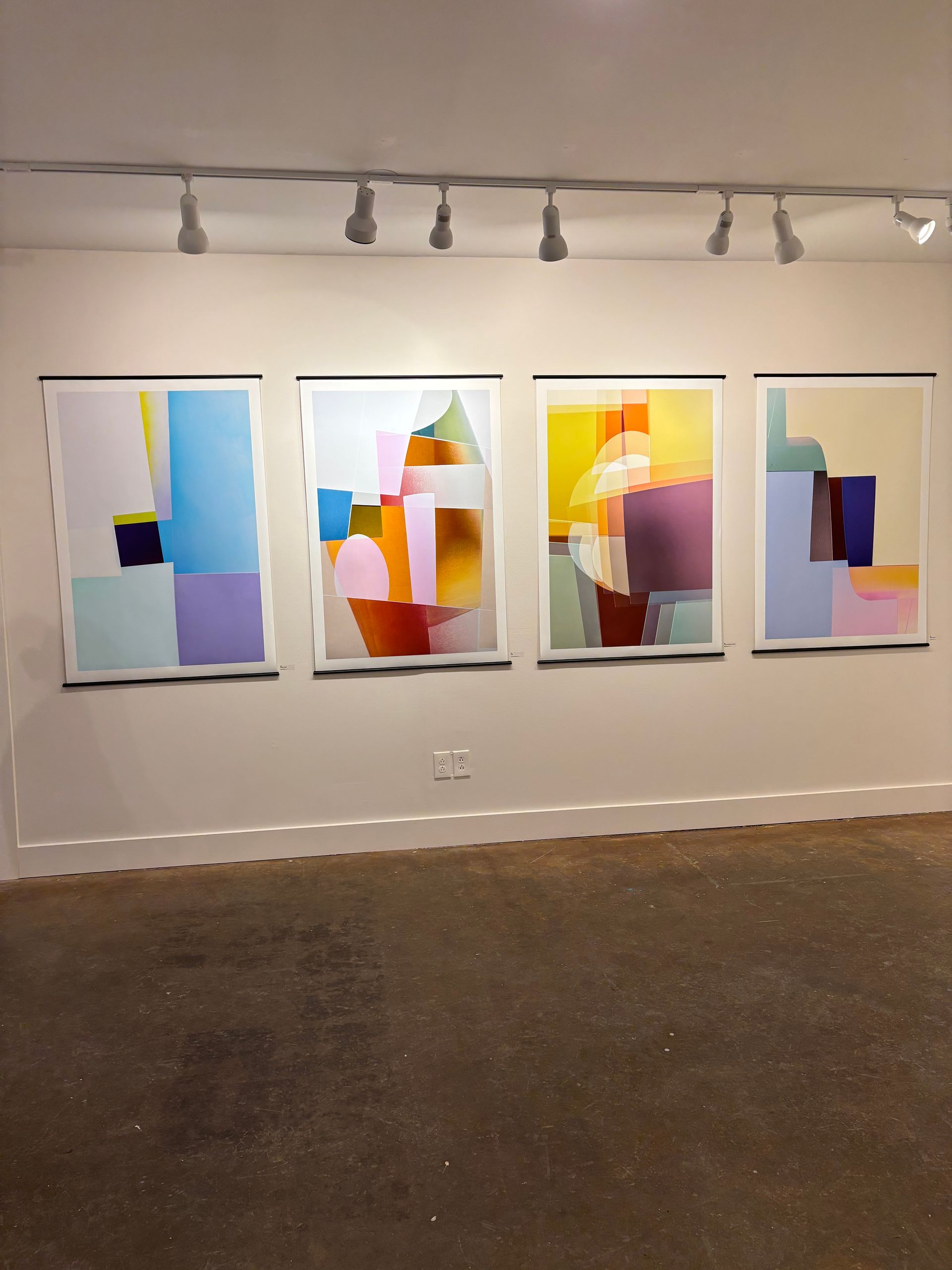 Four abstract colorful paintings displayed in a gallery on a white wall under track lighting