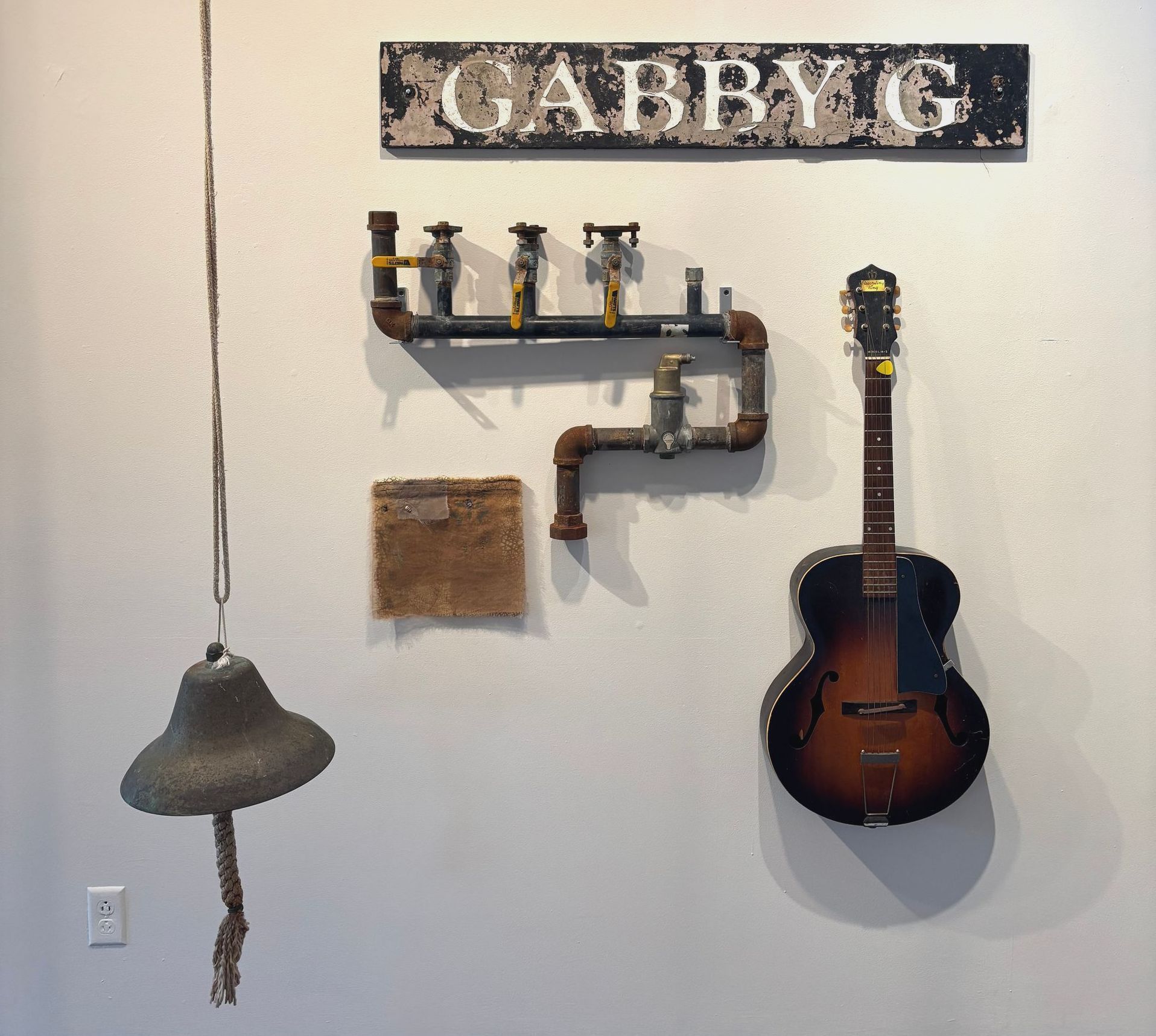 Wall with “GABBY G” sign, metal pipe shelf, hanging bell, and sunburst guitar on display.