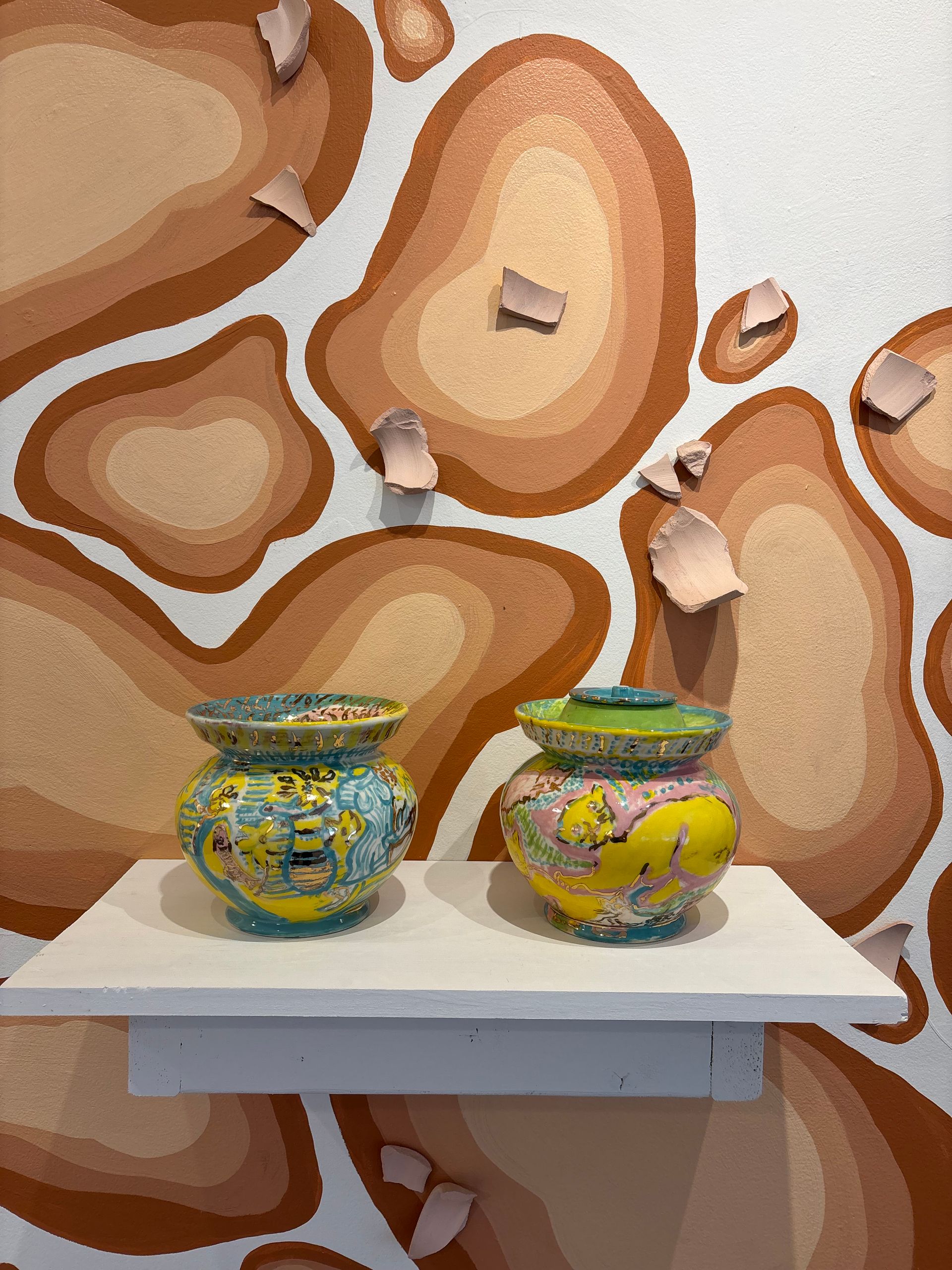 Two colorful decorative jars on a white shelf against a brown abstract mural background