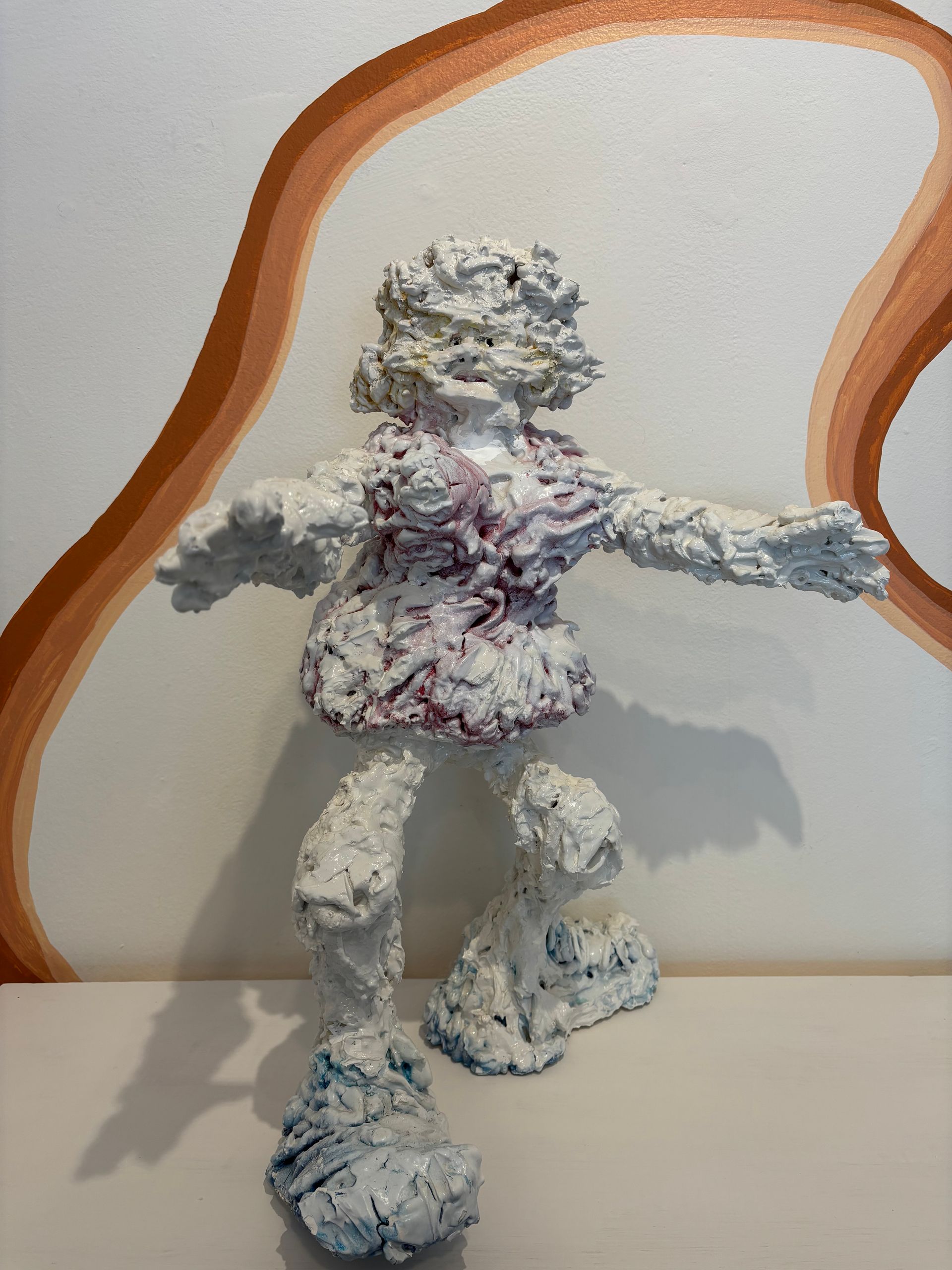 White figurine with arms outstretched, covered in colorful textured paint, against a plain wall.