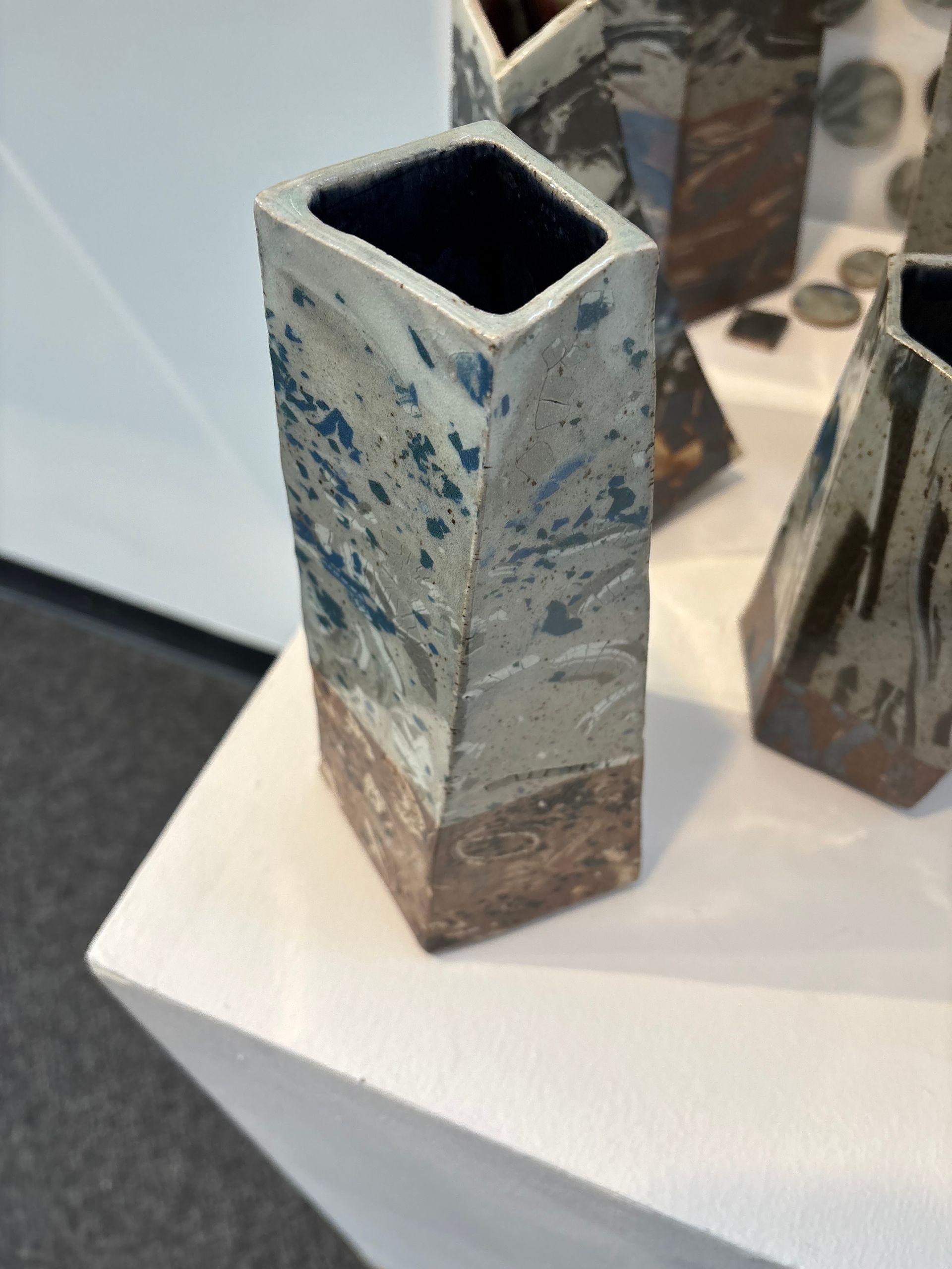 Tall square ceramic vase with blue-gray and brown speckled glaze on a white display table