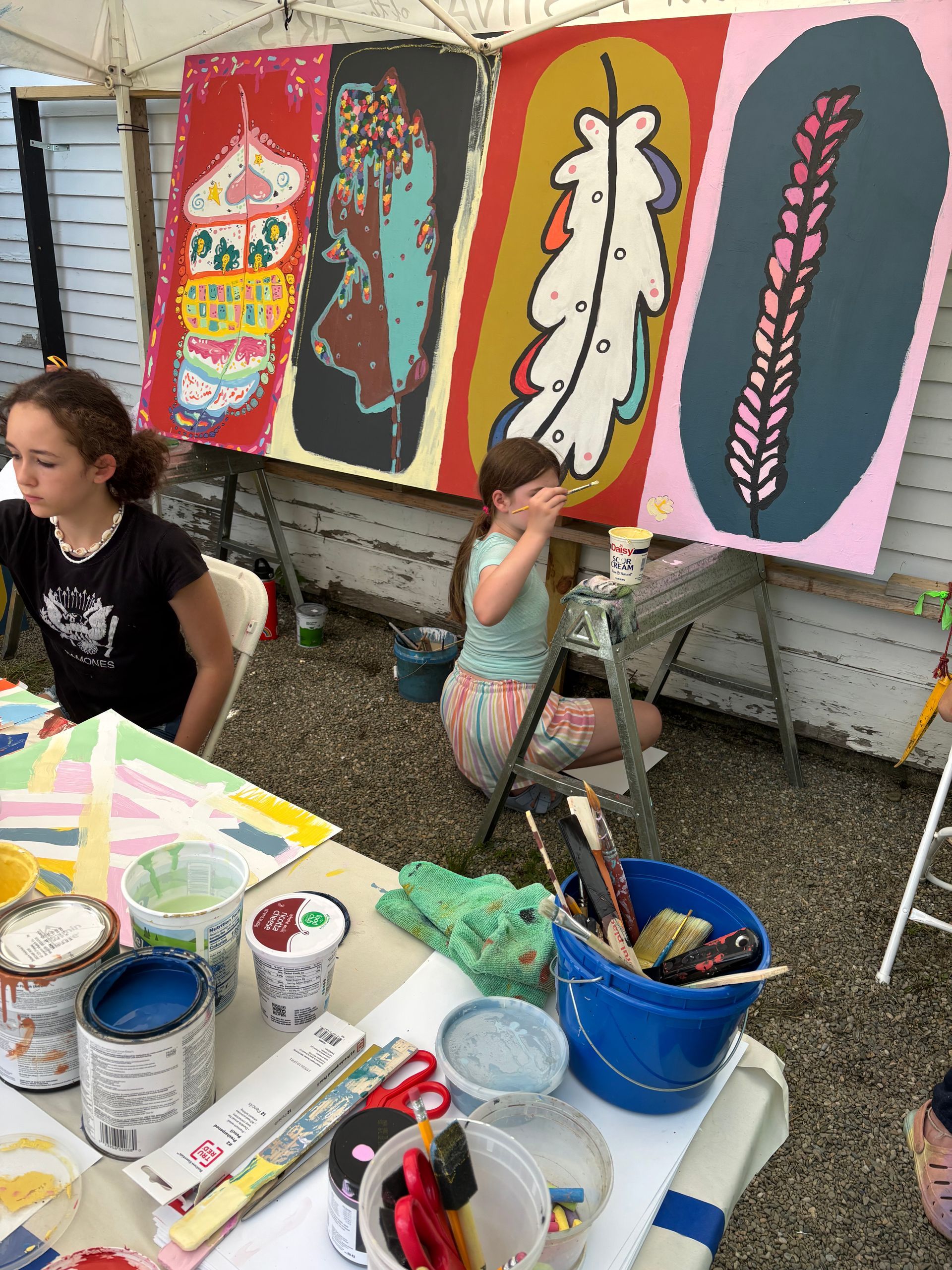Outdoor art station with colorful feather paintings on easels and paint supplies on a table
