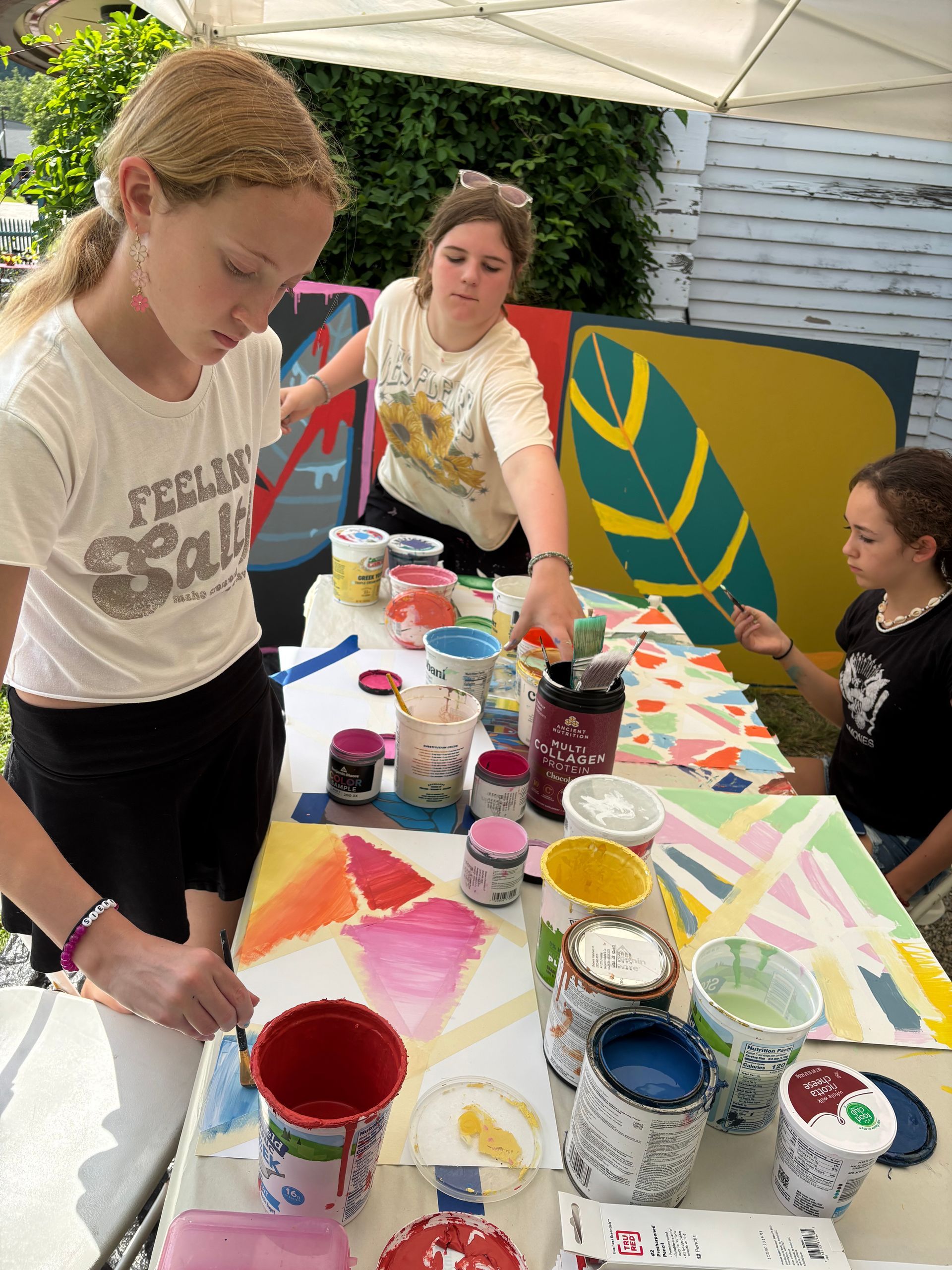 People painting at a table with colorful paint cups under a canopy.