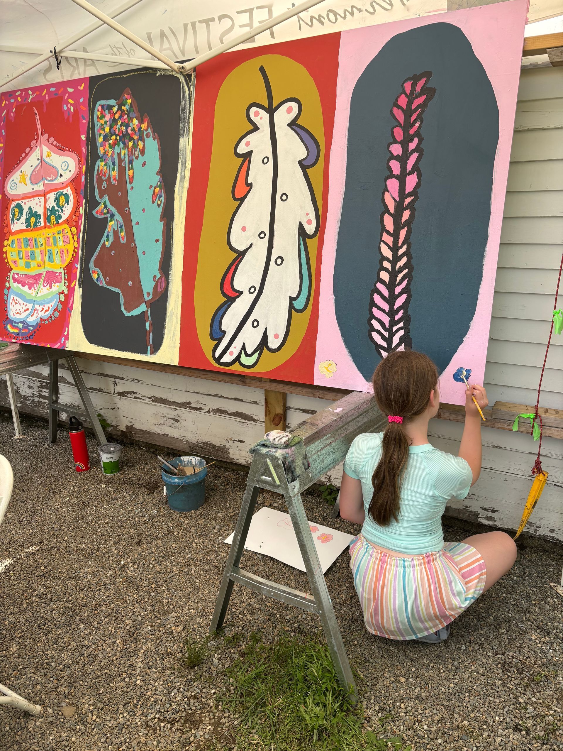 Person painting a colorful mural on a wall outdoors, seated on a step stool amid paint supplies.