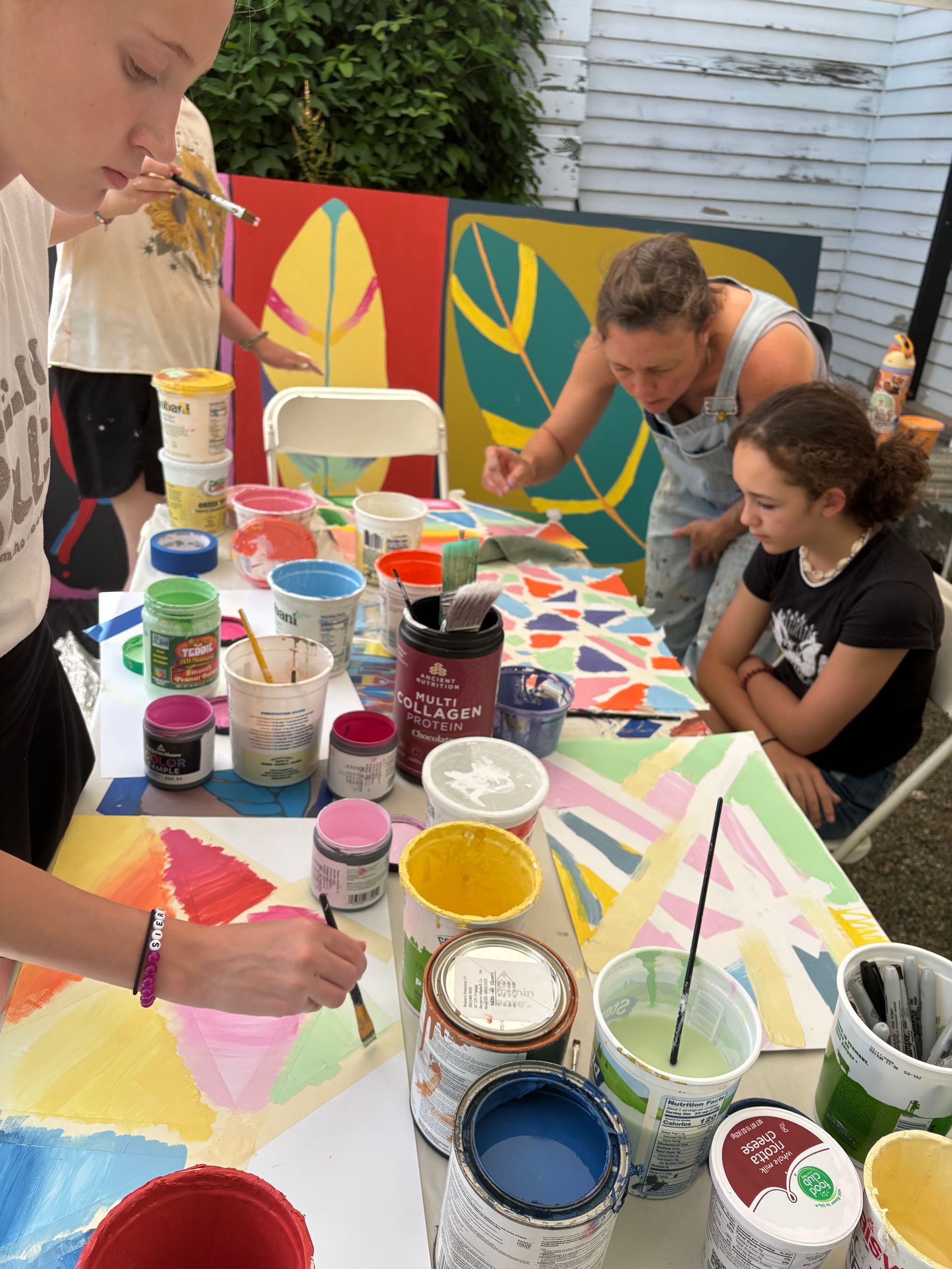 People painting colorful abstract canvases at an outdoor table with open paint jars