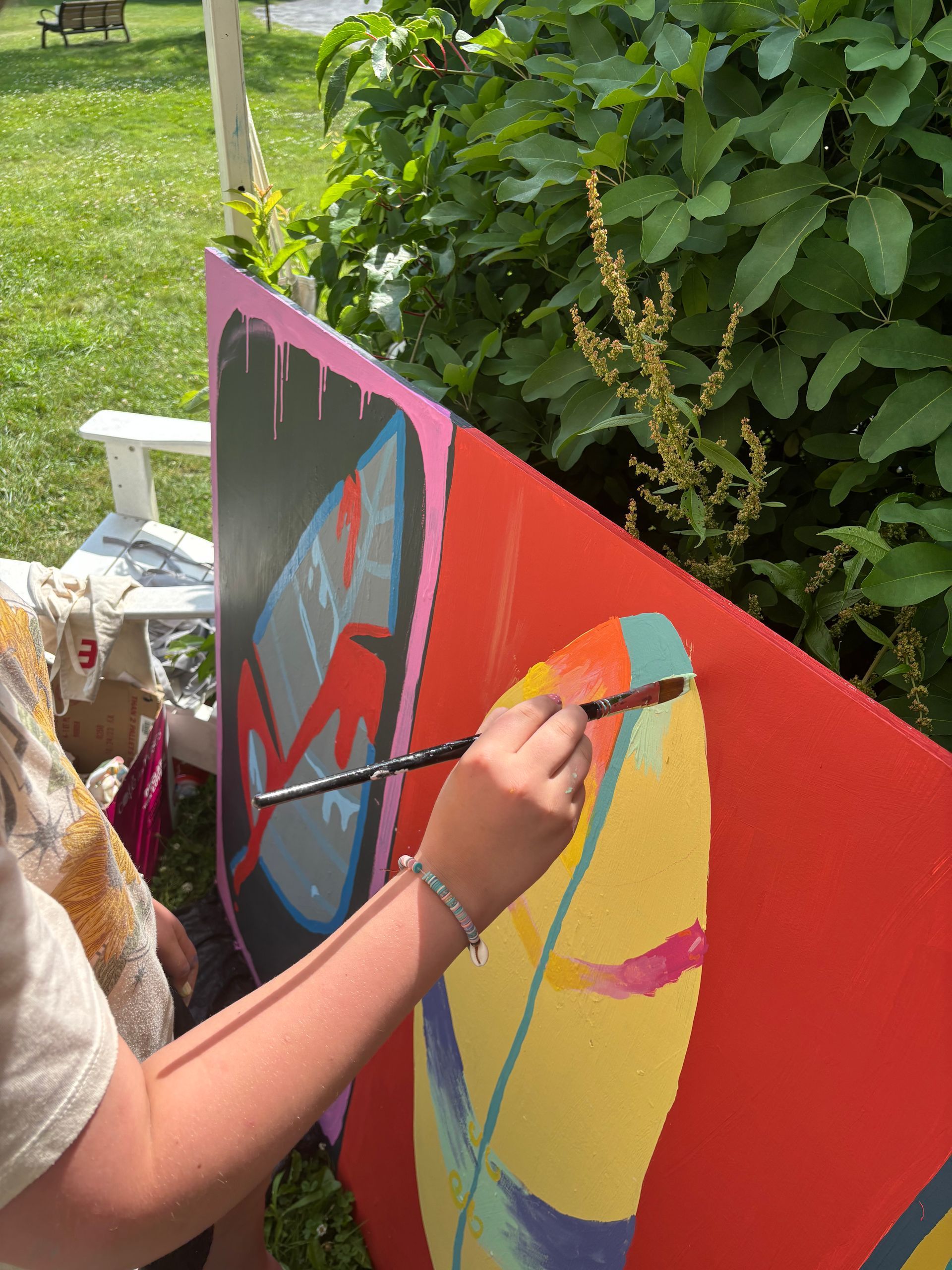Person painting a colorful mural on a red board outdoors beside green bushes