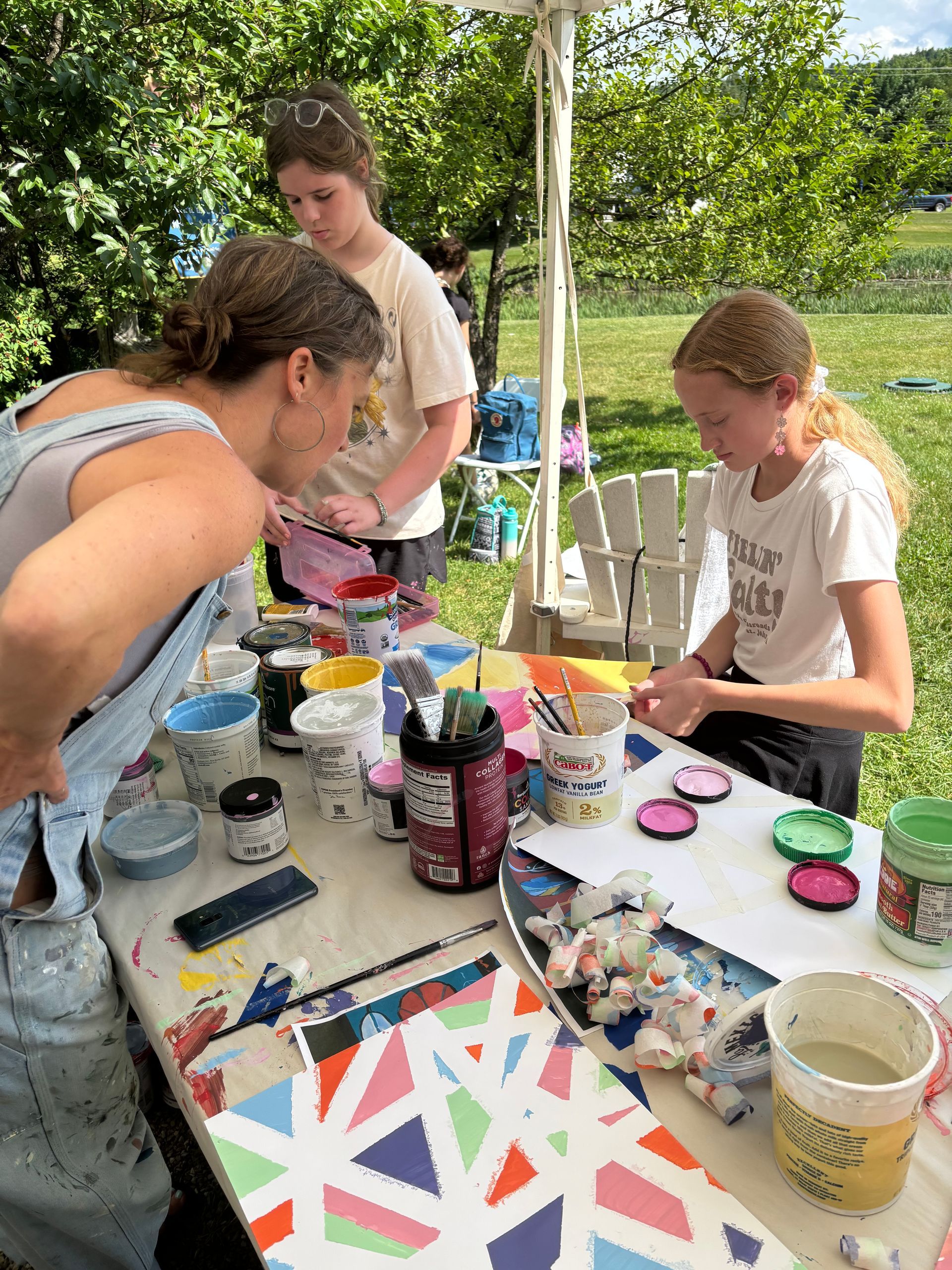 People painting at an outdoor table with colorful art supplies and canvas boards.