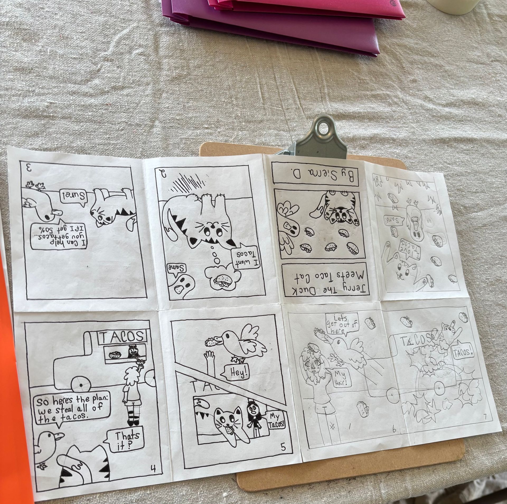 Black-and-white comic pages laid out on a fabric surface, with a clipboard holding one page.