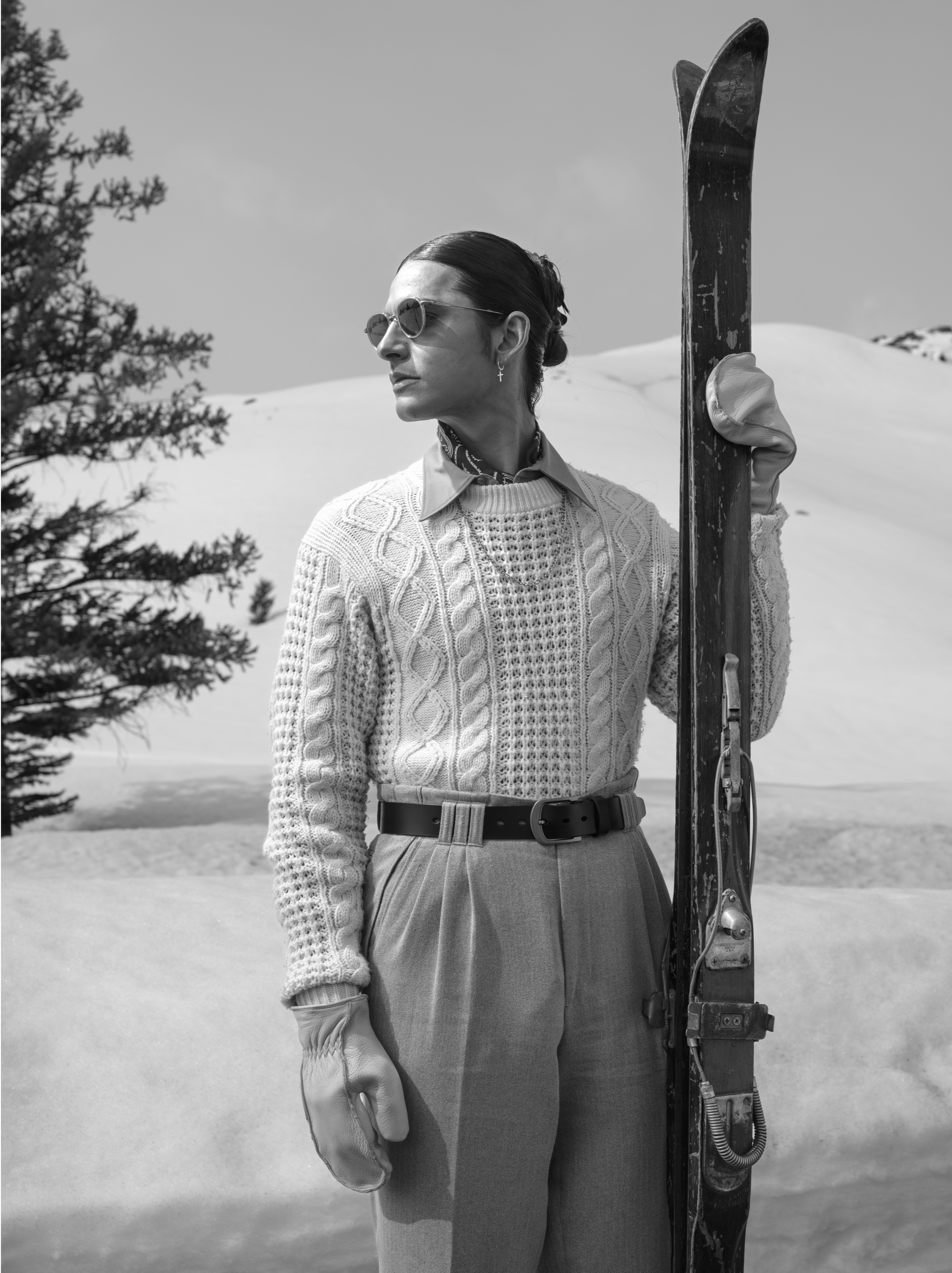Person in sunglasses stands beside skis in a snowy landscape, wearing a patterned sweater and trousers.
