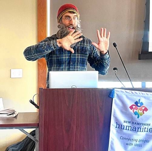 Speaker at a podium gesturing during a presentation, with a laptop and conference banner nearby.
