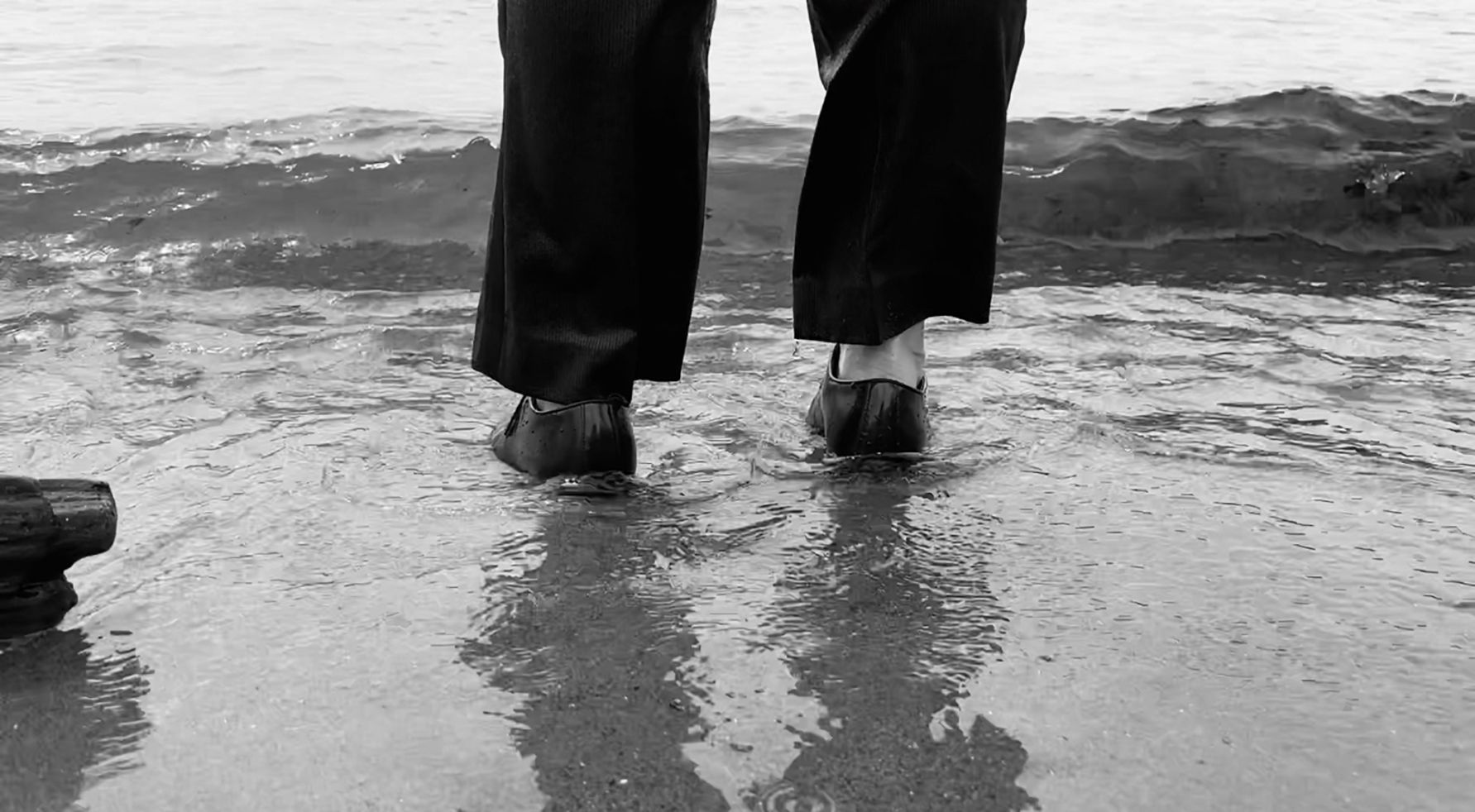 Person standing in shallow surf on a beach, with waves and wet sand reflected below.
