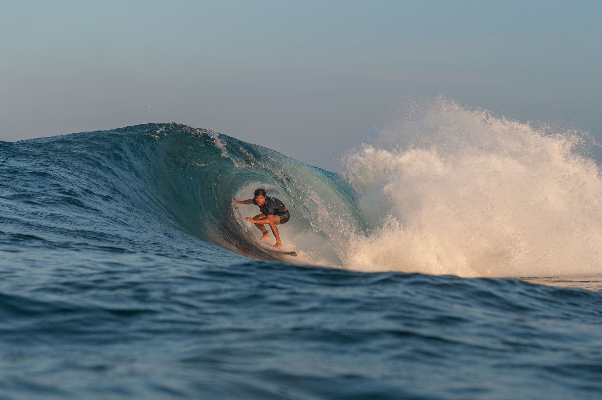 Surfer riding inside a blue wave with white spray at sunset