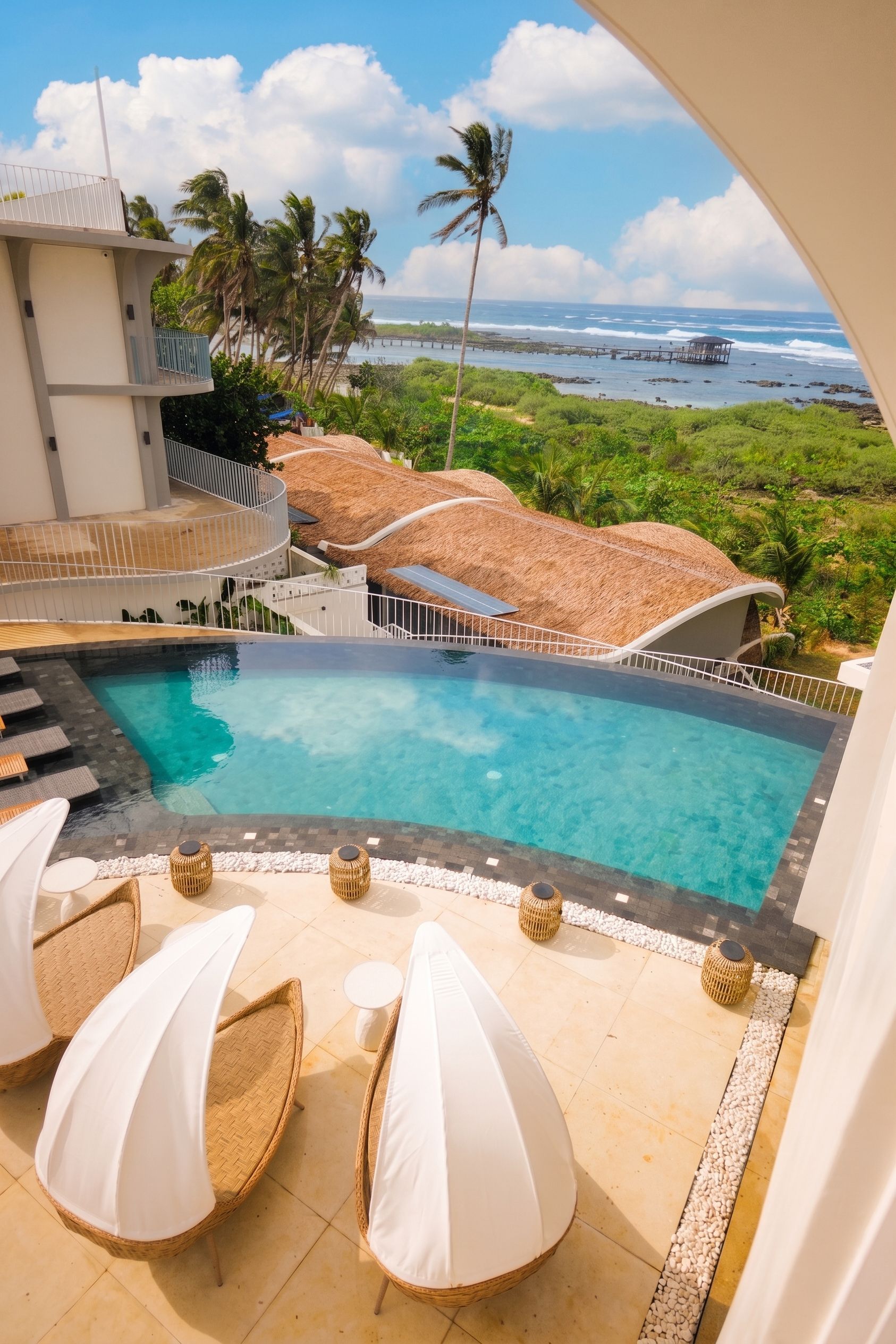 Resort pool overlooking the ocean, with white lounge chairs and palm trees under a bright sky.