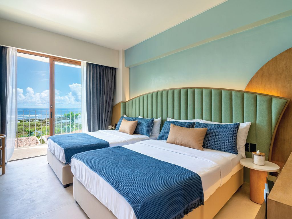 Bright hotel bedroom with teal bed, blue accents, and ocean-view balcony door