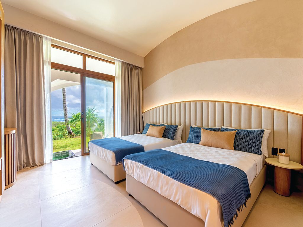 Bright hotel bedroom with twin beds, teal headboard, and ocean view through balcony doors