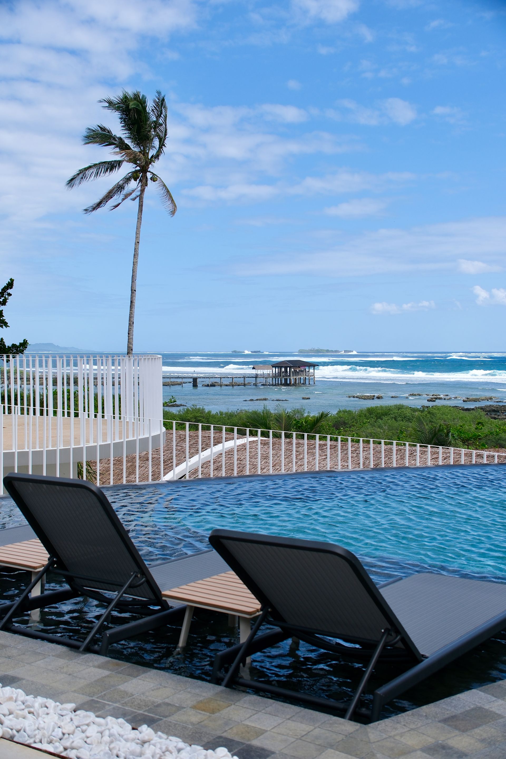Oceanfront pool with two lounge chairs, palm tree, and waves under a blue sky.