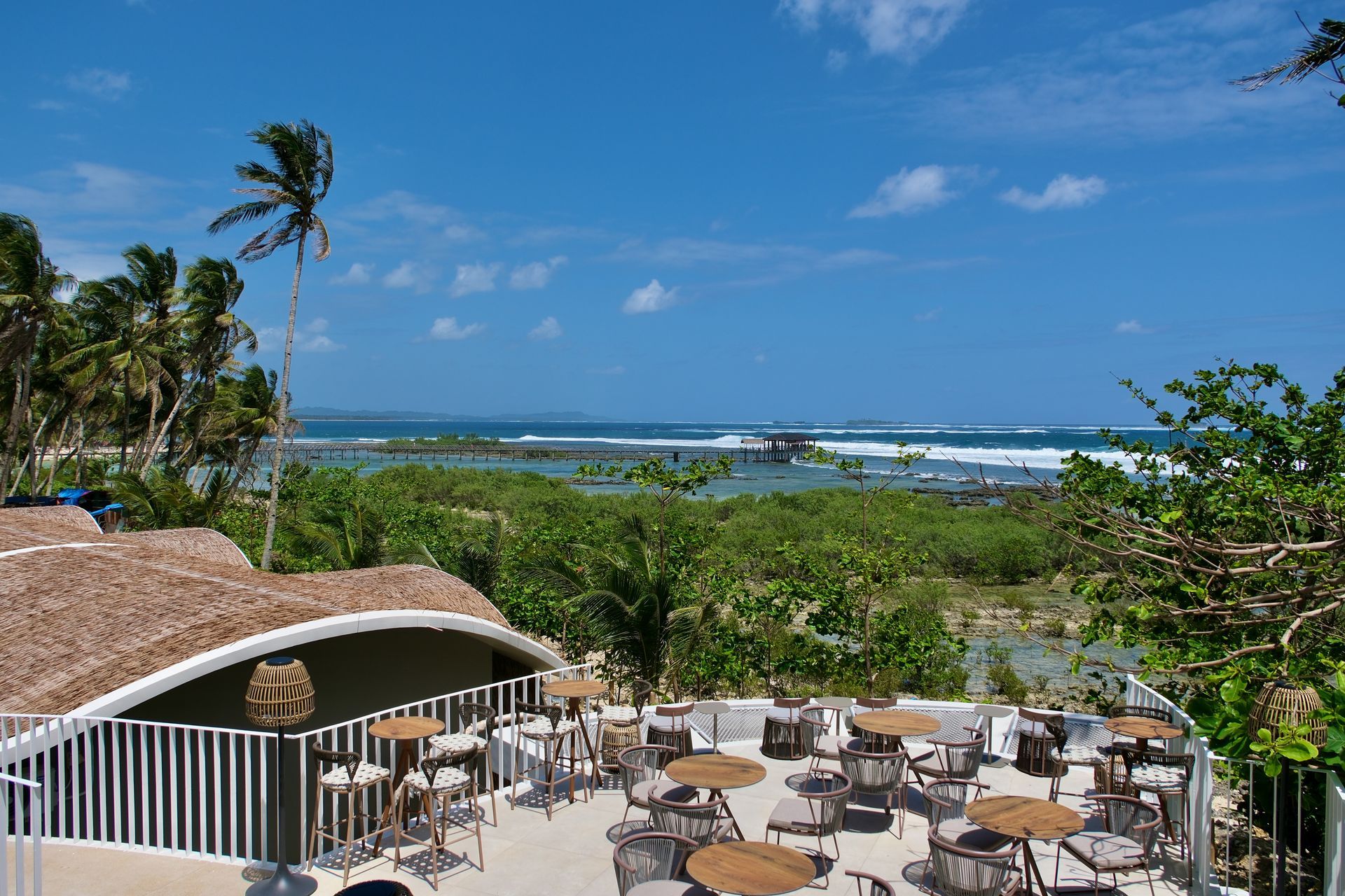 Oceanfront patio with tables and chairs overlooking a blue sea and palm trees under a sunny sky