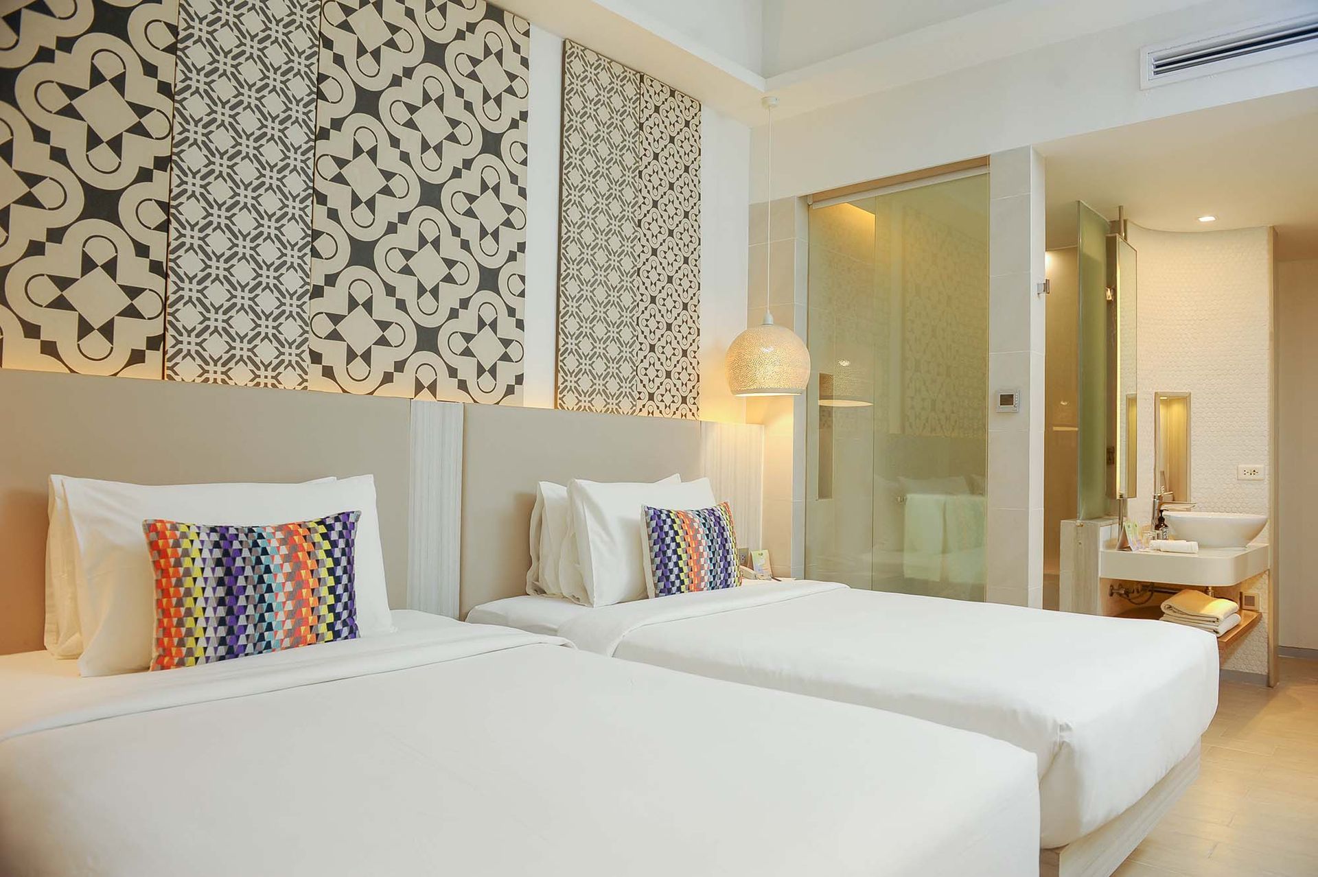 A hotel room with two white beds featuring geometric patterned accent panels on the wall and a modern bathroom in view.