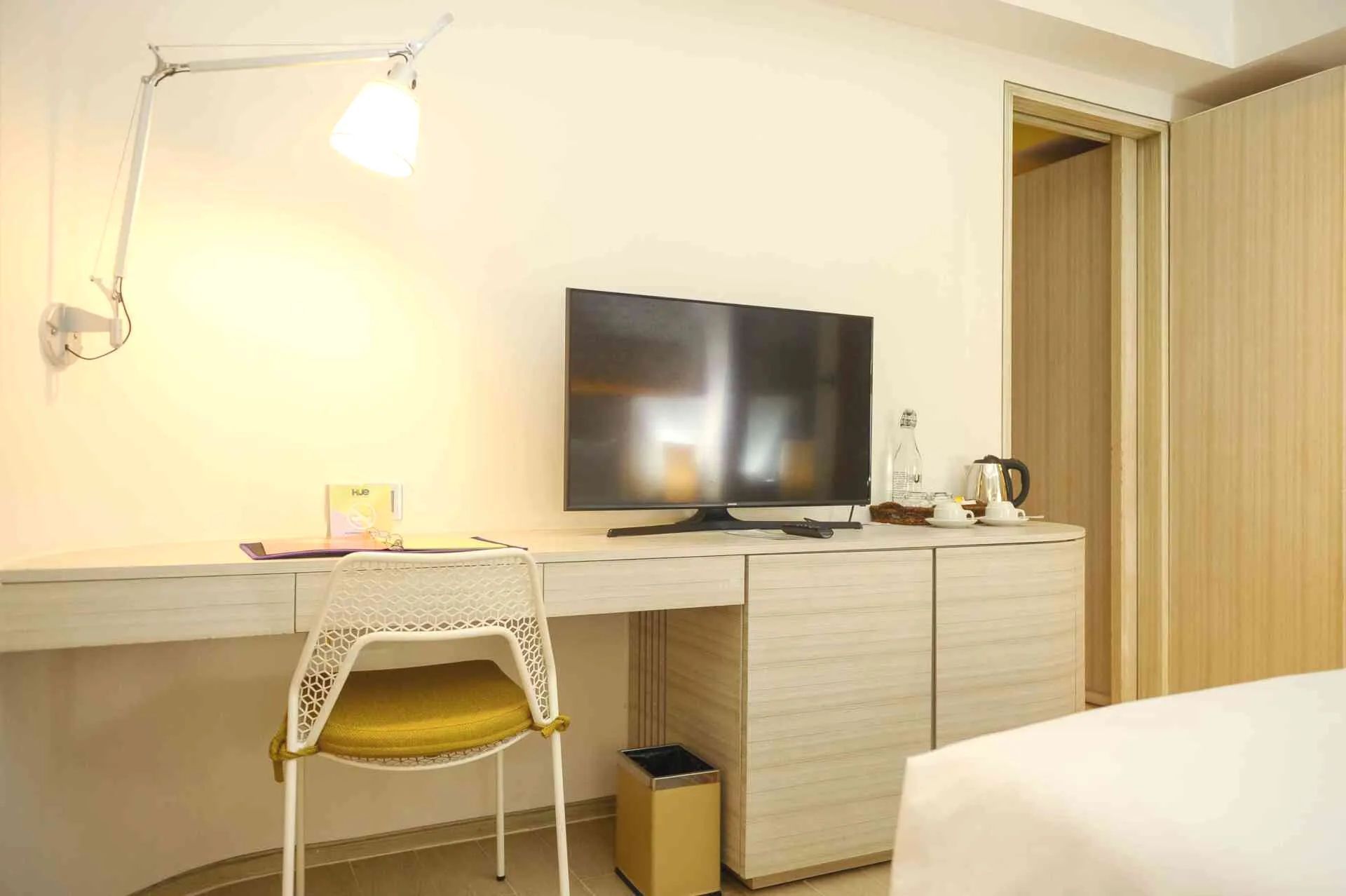 A hotel room with a desk, chair, TV, and lamp, next to a bedroom.