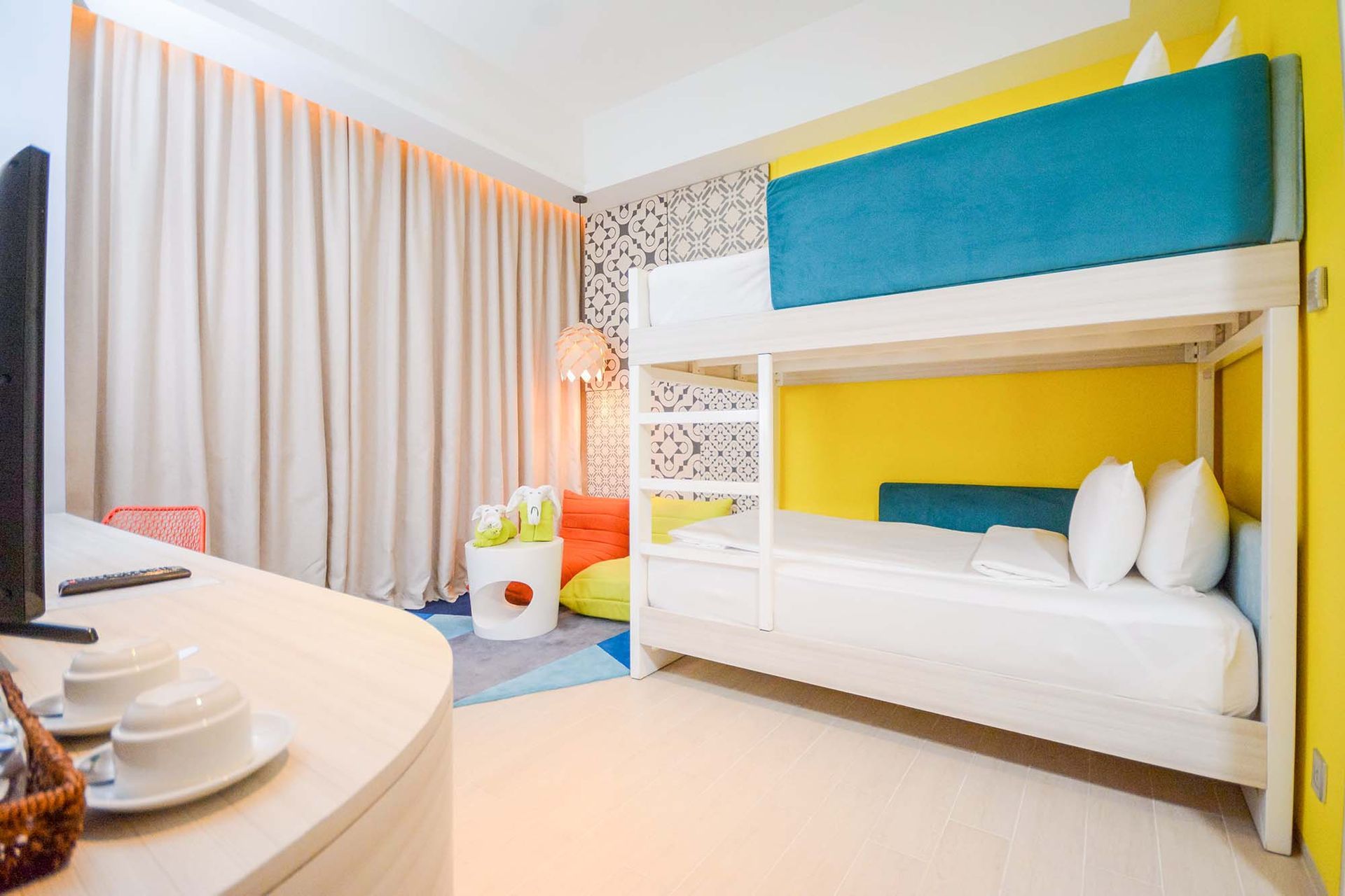 Bright hotel room with bunk beds, white bedding, yellow and teal walls, and a curved desk.