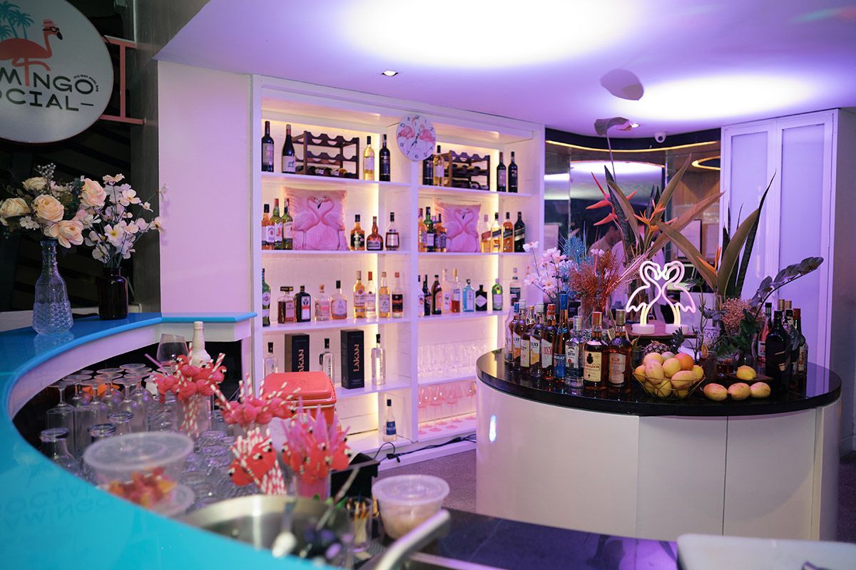 A well-lit bar area features a curved blue countertop, white illuminated shelving stocked with liquor, and tropical decor.