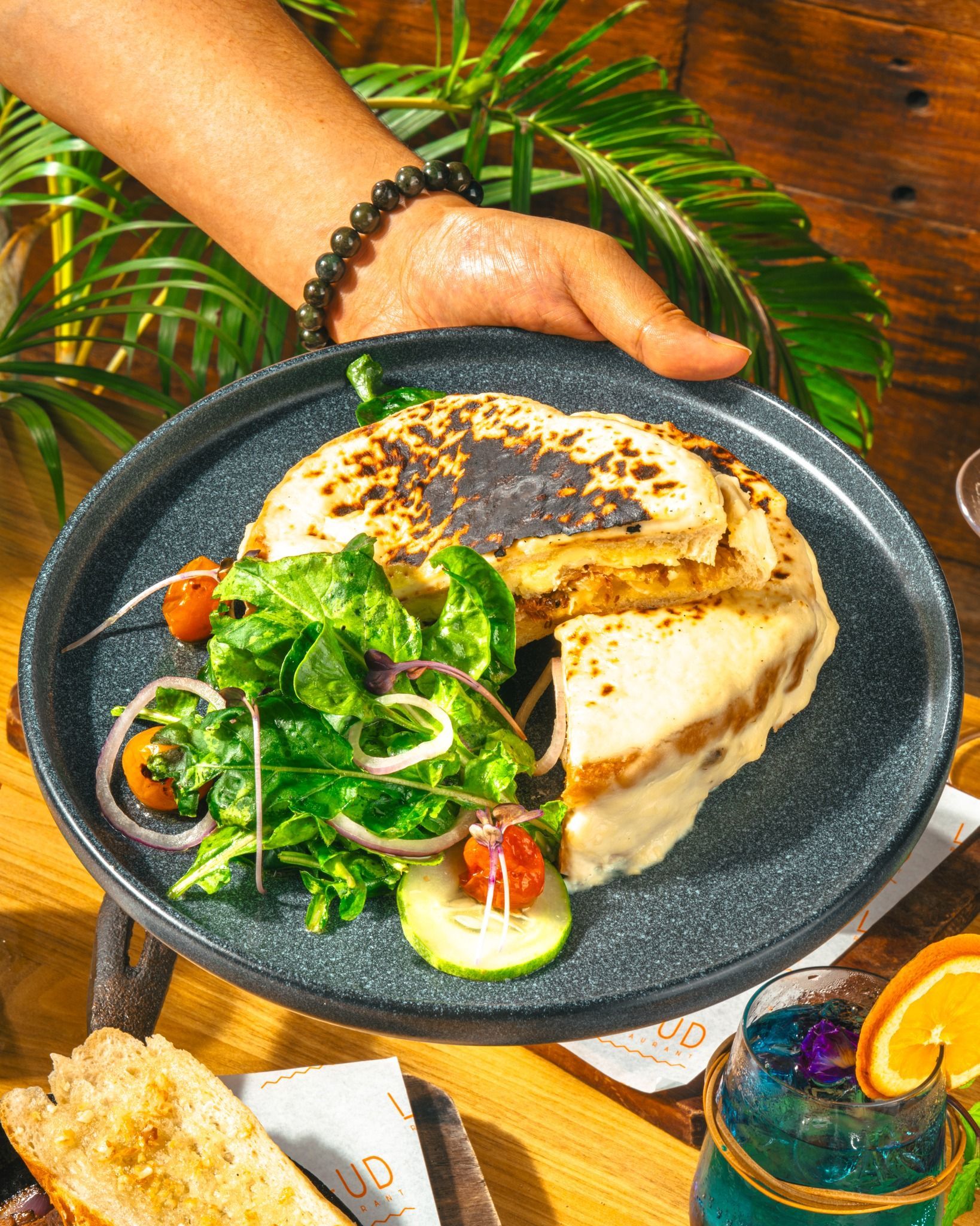 Hand holding a grilled flatbread on a black plate with greens and tomatoes on a wooden table