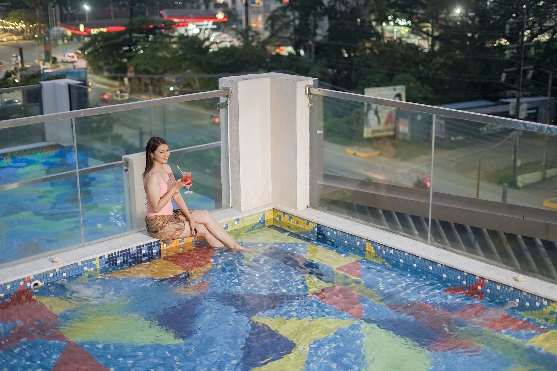 Woman in a pool on a rooftop, holding a drink. Night view of a city in background.