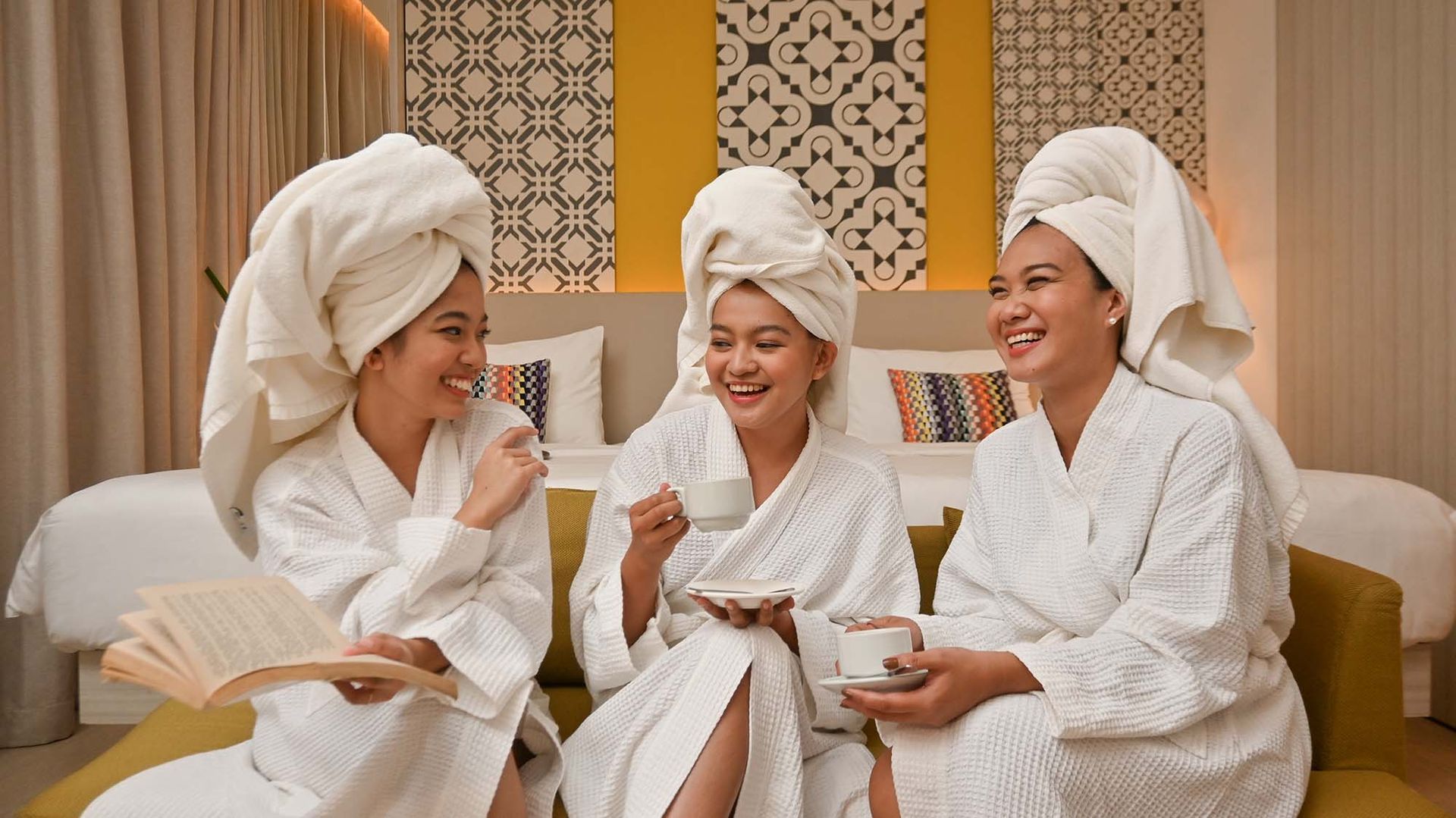 Three people in white robes and towels sit on a bed, smiling and chatting in a cozy spa room.
