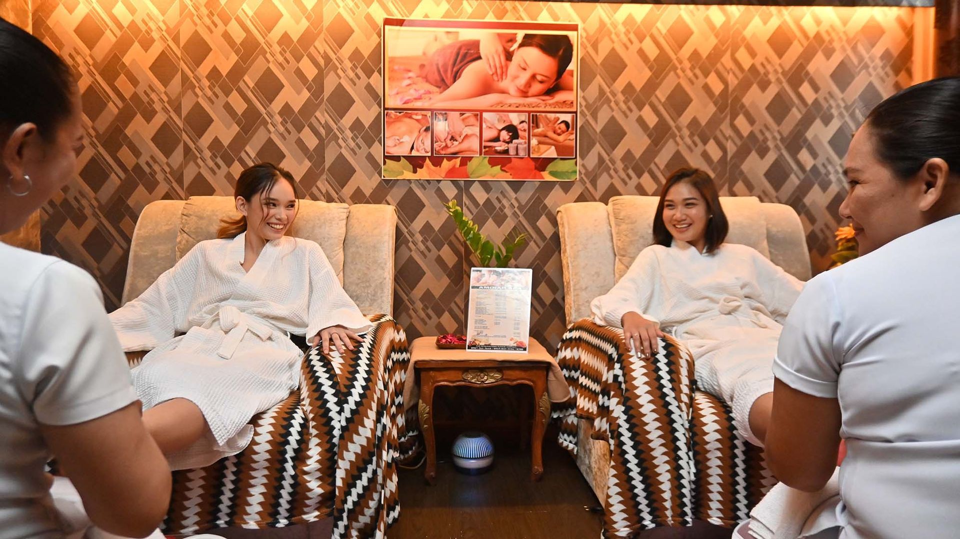 Two people in white robes receiving spa treatments in a warm, patterned massage room.