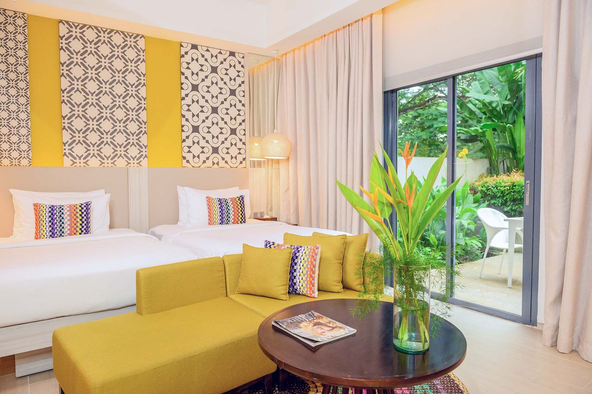 Bright living room with yellow sofa, white daybed, patterned wall art, and sliding glass doors to greenery