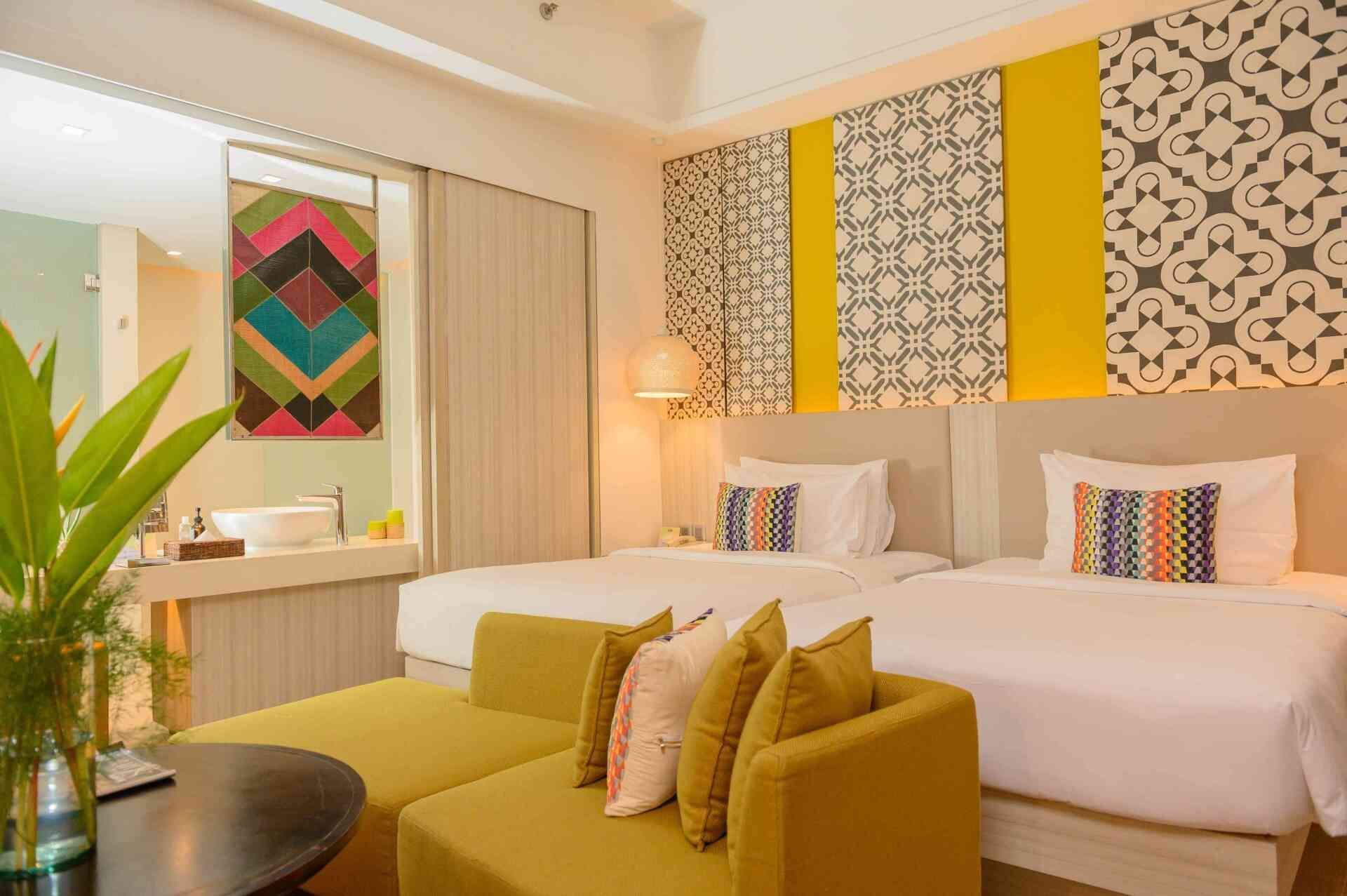 Bright hotel room with twin beds, colorful geometric wall art, mustard sofa, and large potted plant