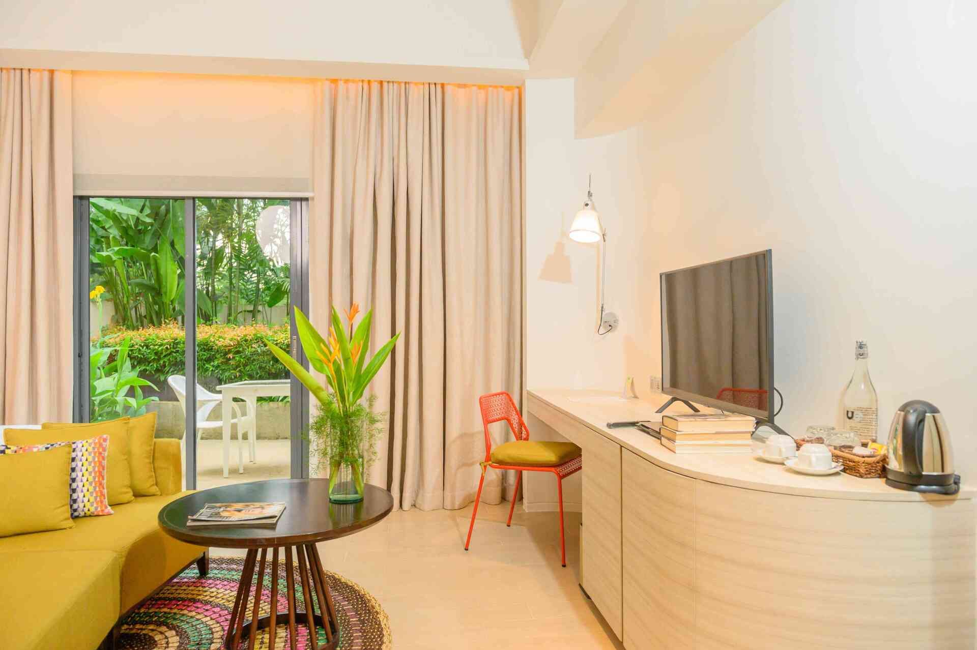 Yellow-themed hotel room with a sofa, desk, TV, and sliding glass door to a patio with greenery.