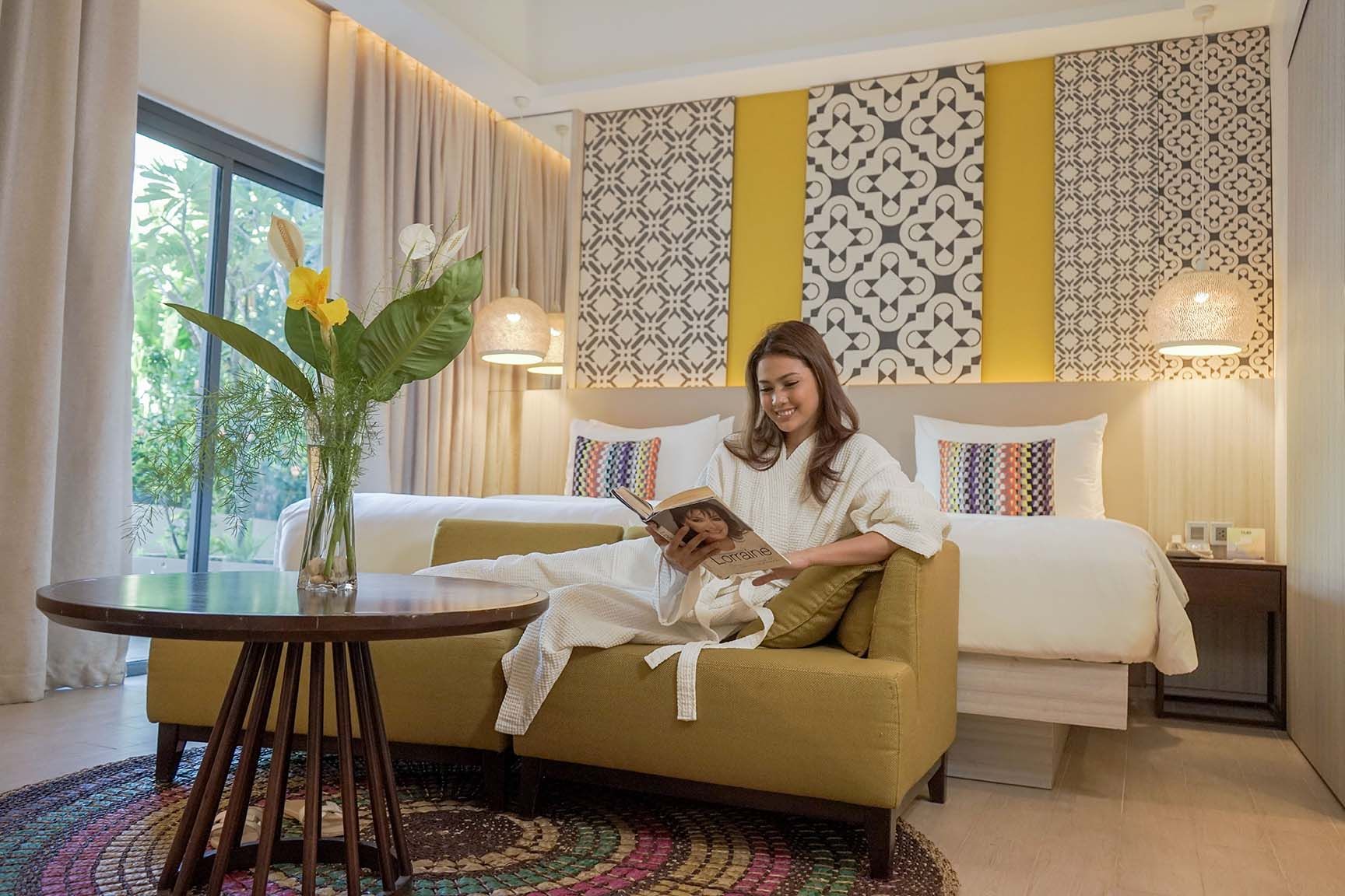 A person in a white robe sits on a yellow sofa in a hotel room, reading a book, with a round table and plants in front.