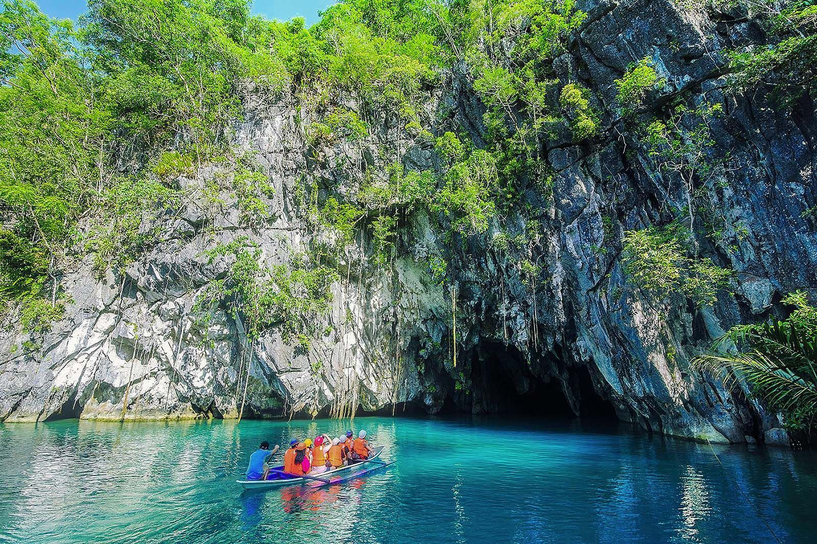 Boat with tourists enters a cave with turquoise water; cliffs are covered in lush green vegetation.