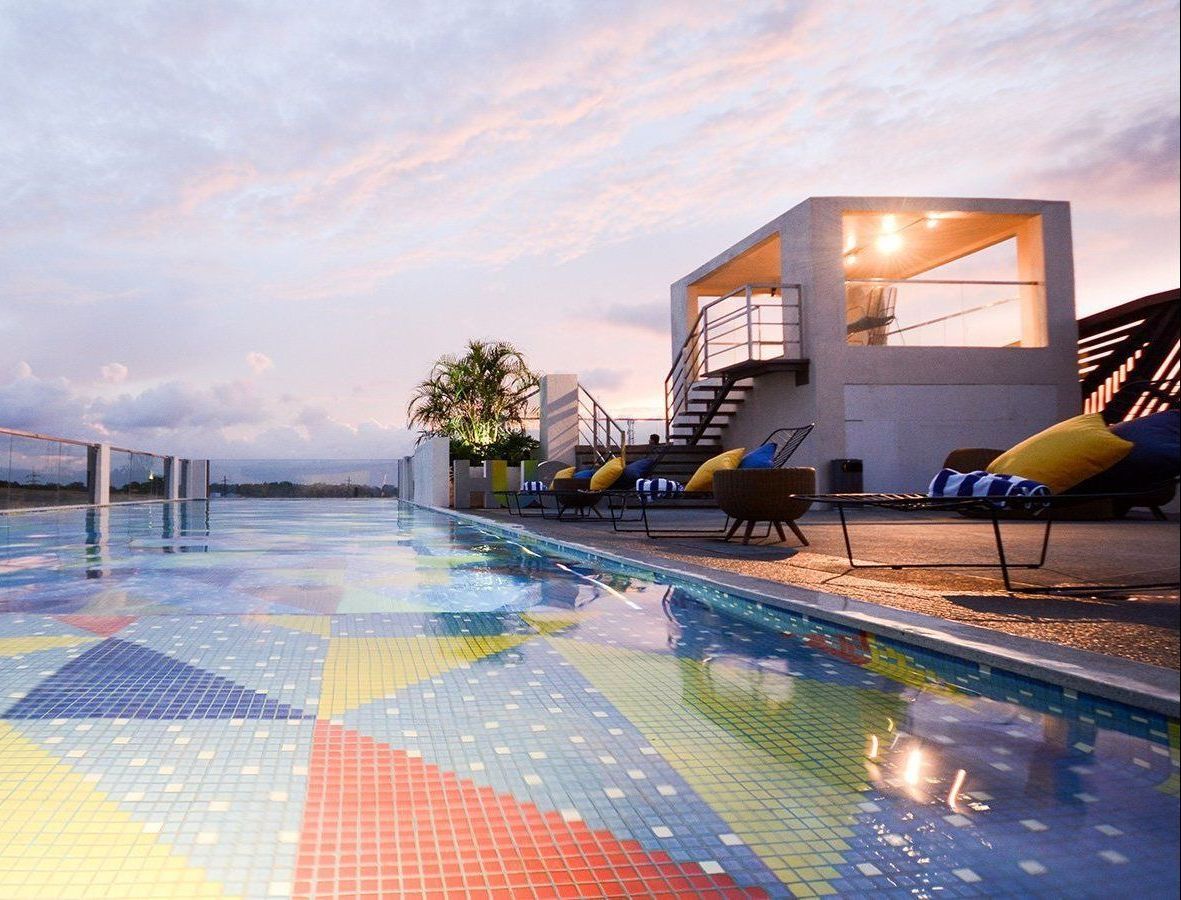 Rooftop pool with colorful mosaic tiles, lounge chairs, and modern building under a cloudy sky.