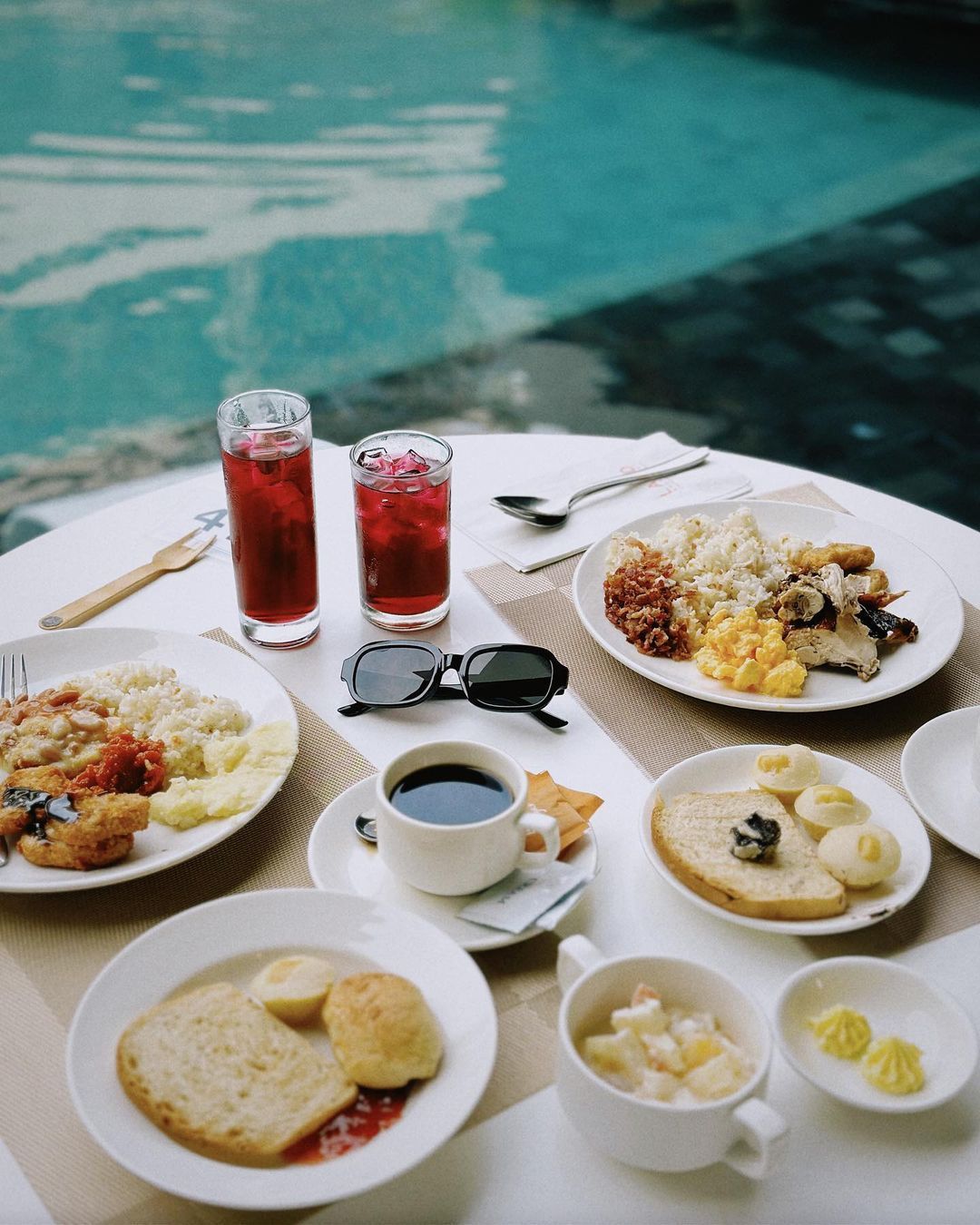 A breakfast spread with plates of food, coffee, and red drinks on a table overlooking a blue swimming pool.