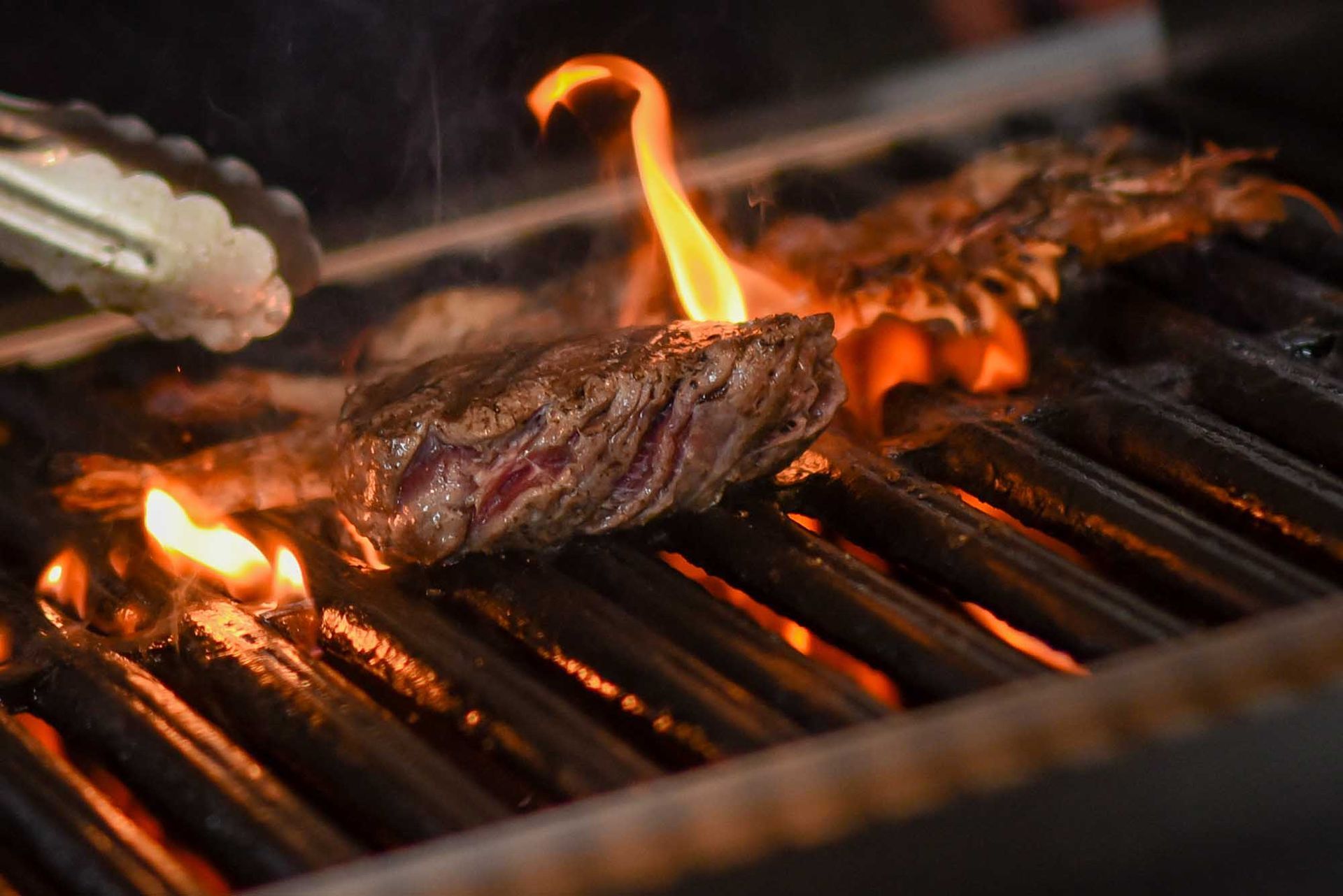 Steak grilling over open flames on a barbecue grate, with tongs nearby