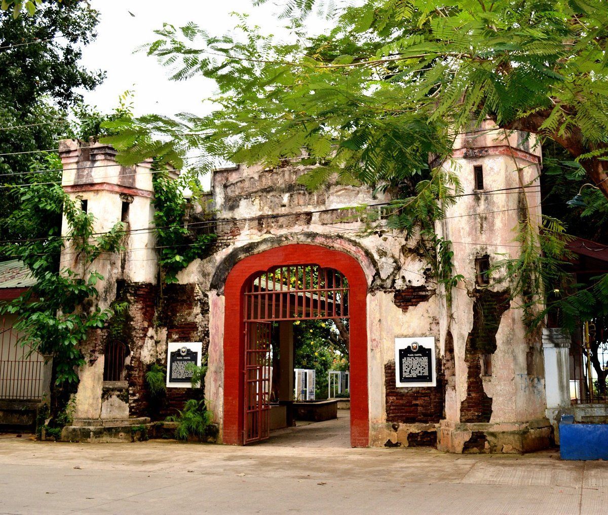 Old stone entrance gate with a red arch, flanked by white pillars and surrounded by trees.