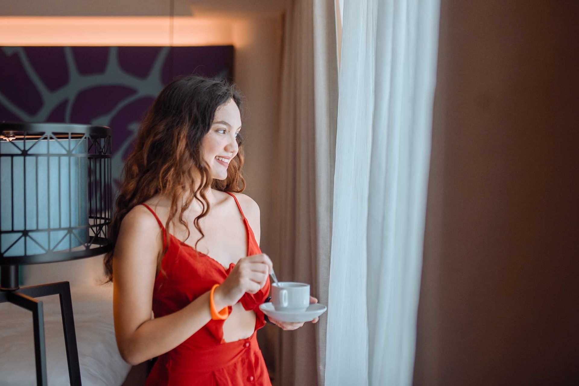 Woman in red dress with coffee cup, looking out window, smiling in a hotel room.