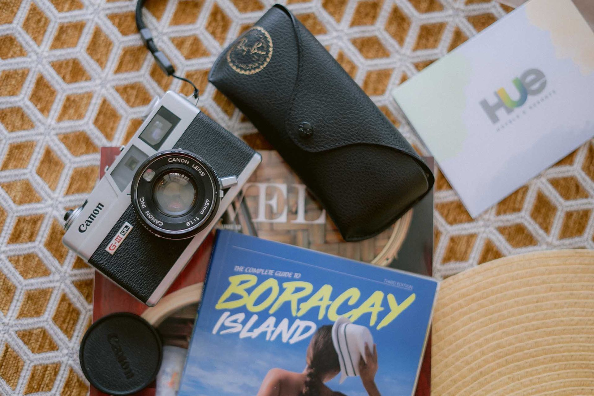 Travel flat lay with a camera, Boracay Island guidebook, sunglasses case, and a white card on a patterned cloth.