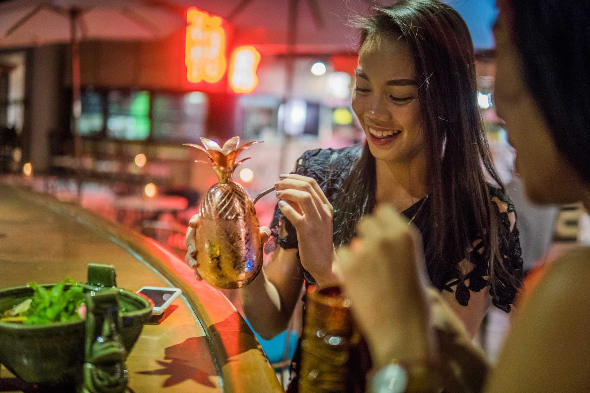Friends dining at an outdoor restaurant, smiling over a hookah lit by warm neon lights.