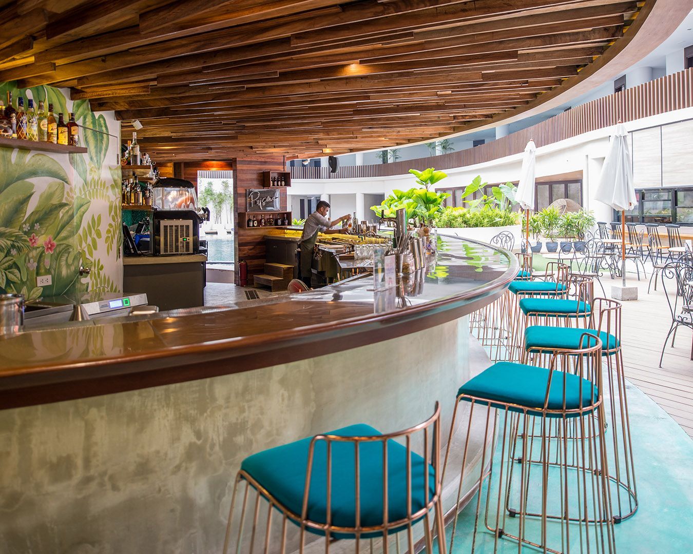 A curved bar with teal stools and a wooden slat ceiling, set in an open-air area with a tropical mural on the wall.