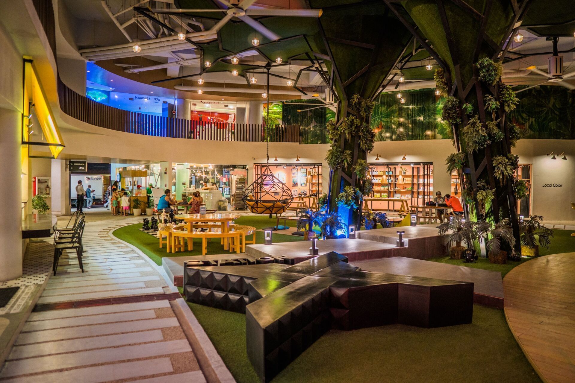 A modern indoor atrium of Station  featuring artificial trees, tiered seating, grass-like rugs, and warm string lighting.
