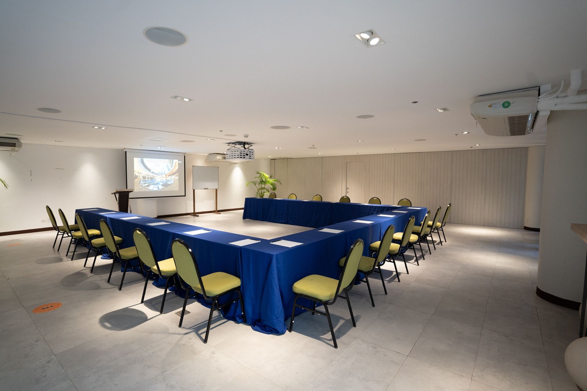 Conference room with U-shaped blue tables, yellow chairs, projector screen, and overhead lighting.