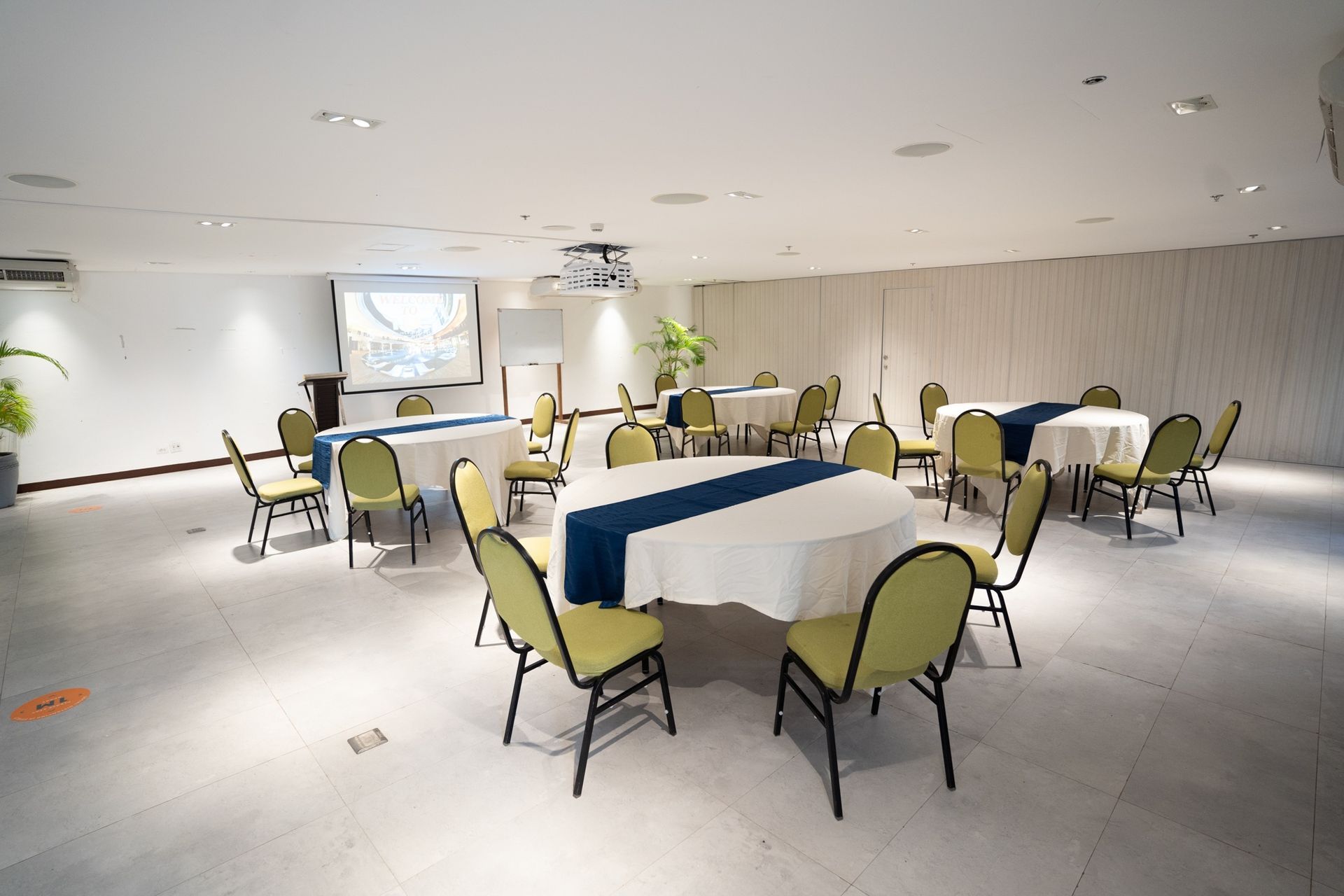 Banquet room with round tables, blue tablecloths, and green chairs under bright ceiling lights.