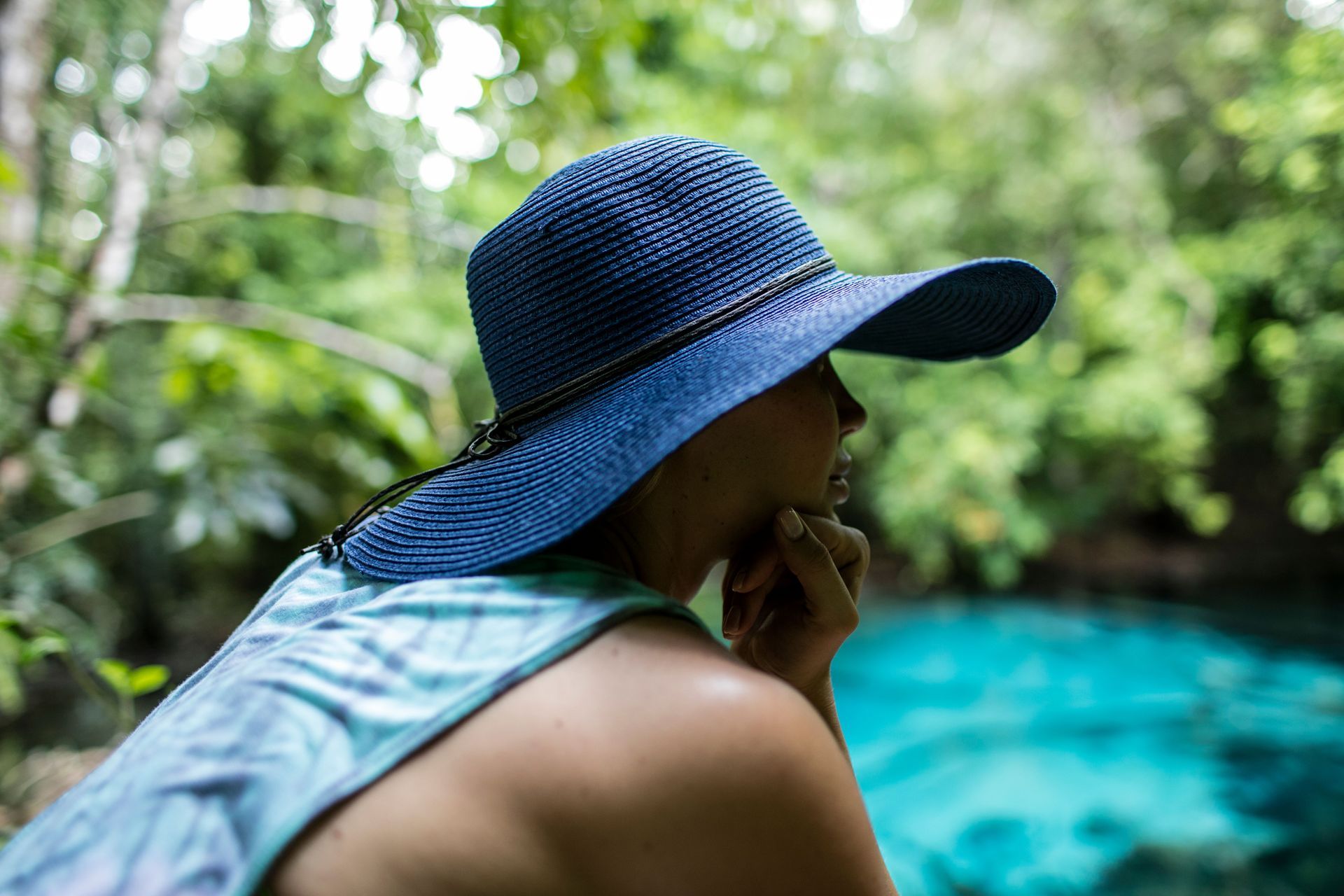 Person in a blue sunhat relaxing by a turquoise pool surrounded by lush greenery