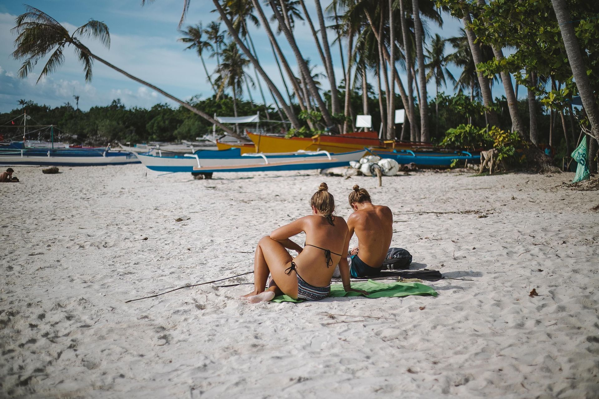 Two people sit on a sandy beach with palm trees and boats in the background.