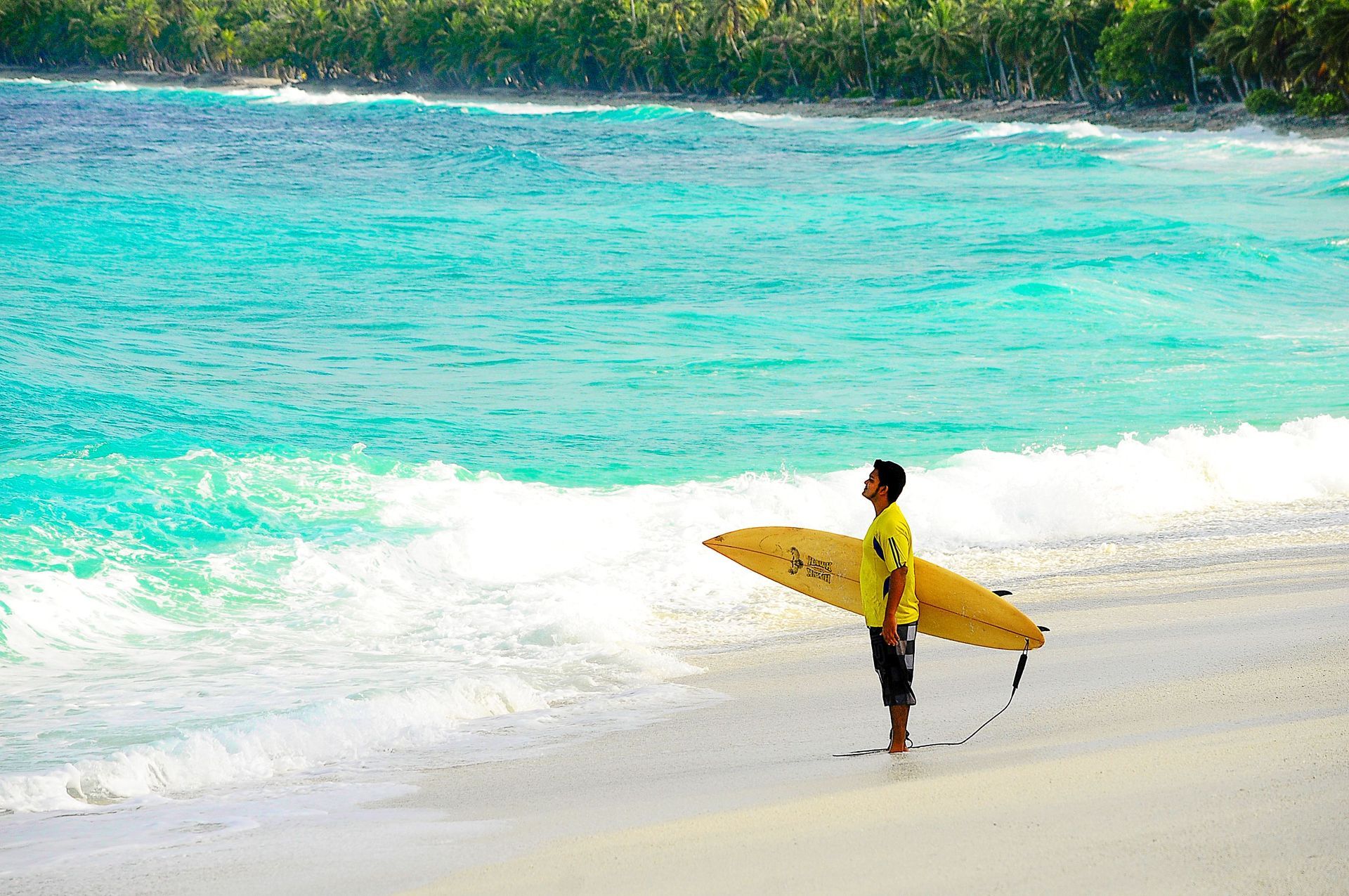 Surfer holding a yellow surfboard on a sandy beach beside turquoise waves