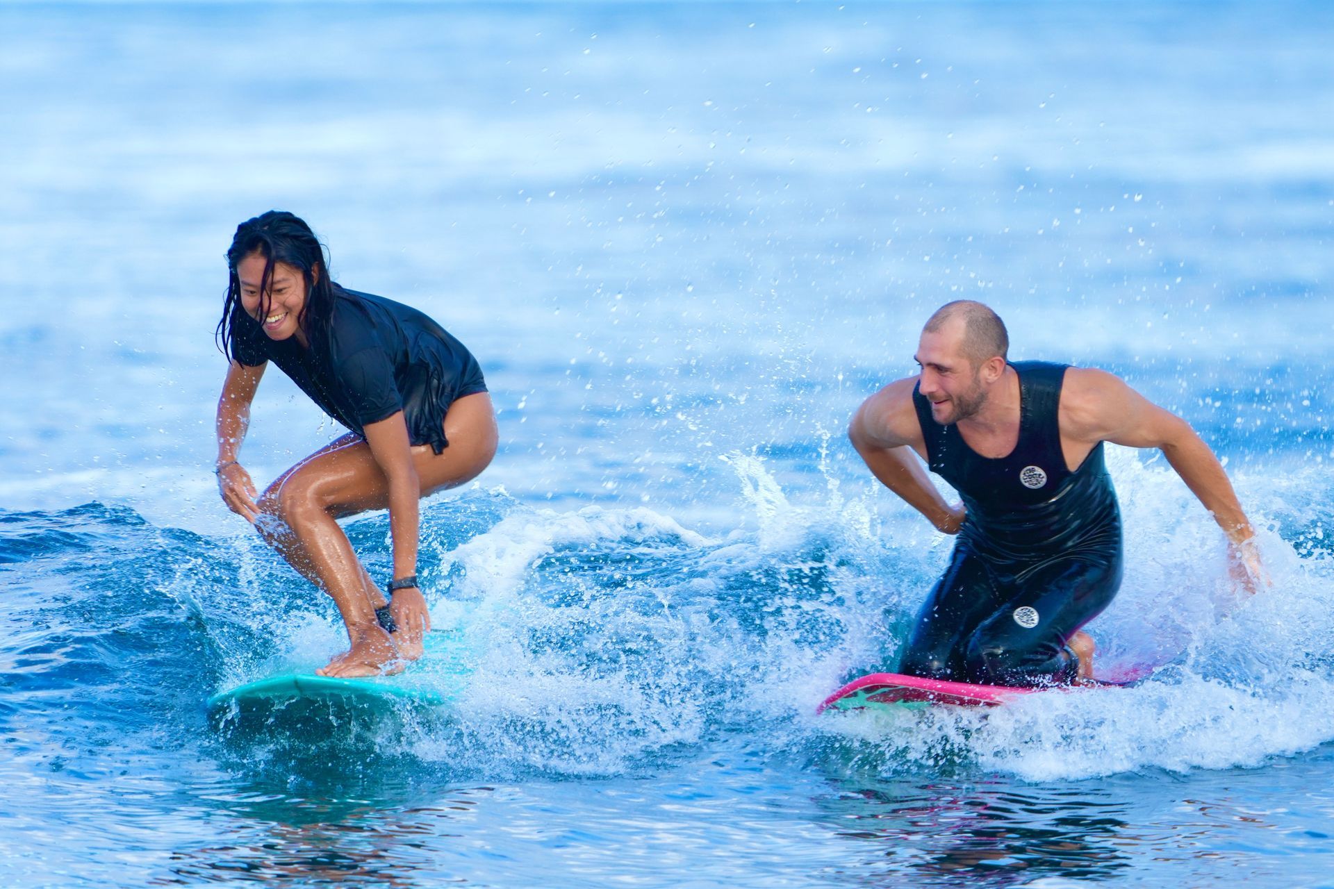 Two surfers crouch on boards riding small waves in the ocean, splashing water.